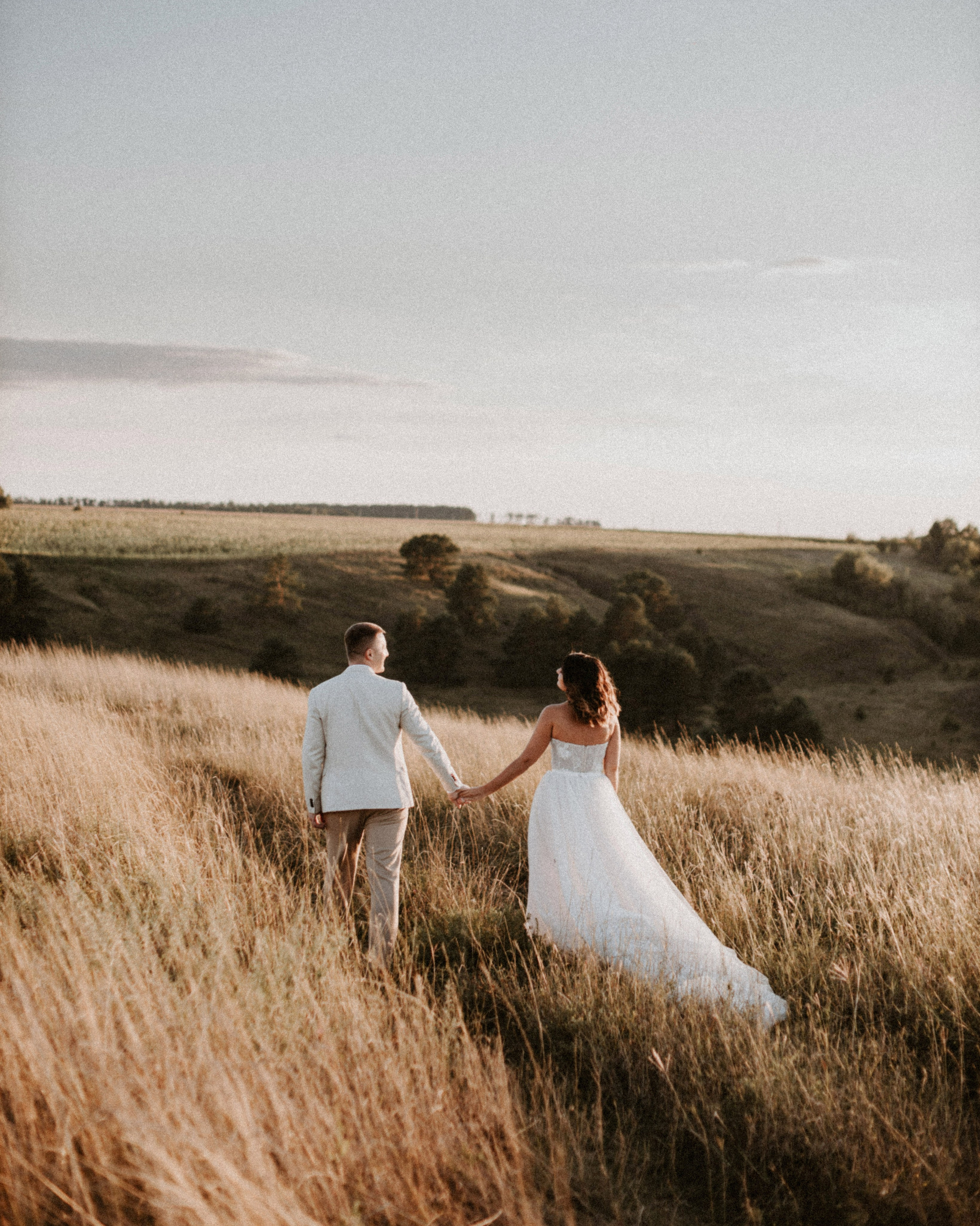 “Una coppia cammina mano nella mano tra l'erba alta e dorata di un campo pittoresco. La sposa, in un fluente abito bianco, fa strada, mentre lo sposo, vestito con un abito chiaro, la segue. Lo sfondo è costituito da colline ondulate, immerse nella luce morbida e calda del sole al tramonto, che crea un'atmosfera serena e romantica”.