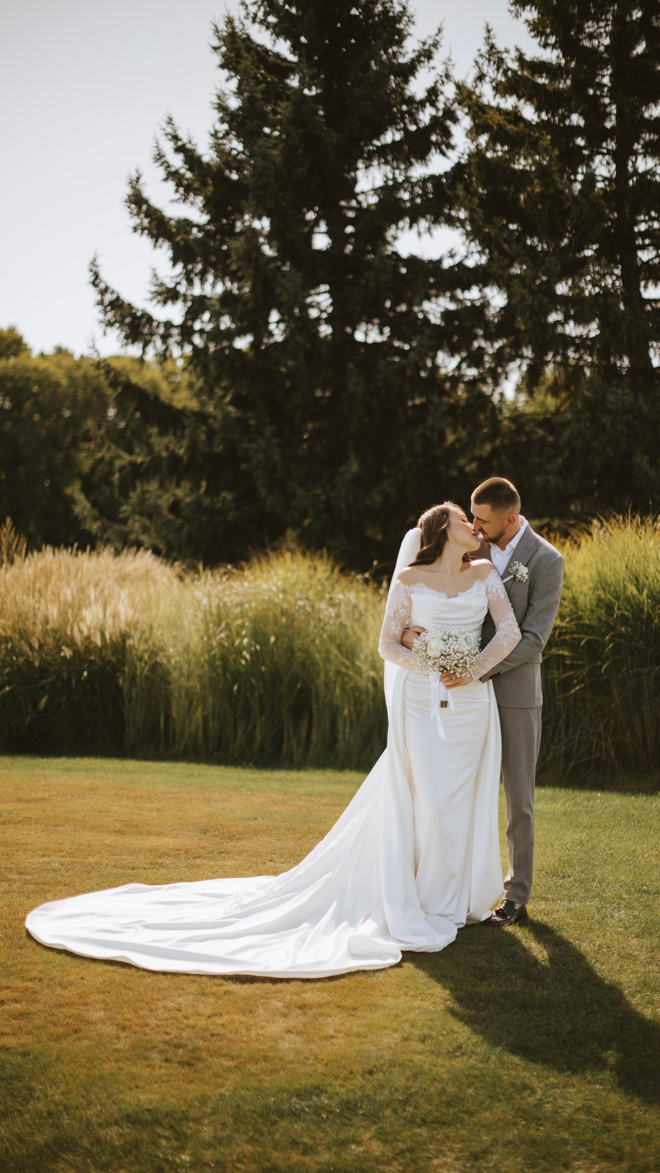 “Una coppia si trova in un ambiente verde e lussureggiante all'aperto e si scambia un tenero bacio. La sposa indossa un abito da sposa di pizzo a maniche lunghe con uno strascico fluente e tiene in mano un piccolo bouquet di fiori bianchi. Lo sposo, in abito grigio chiaro, la abbraccia dolcemente. Alti alberi e campi erbosi li circondano, creando un'atmosfera serena e romantica”.