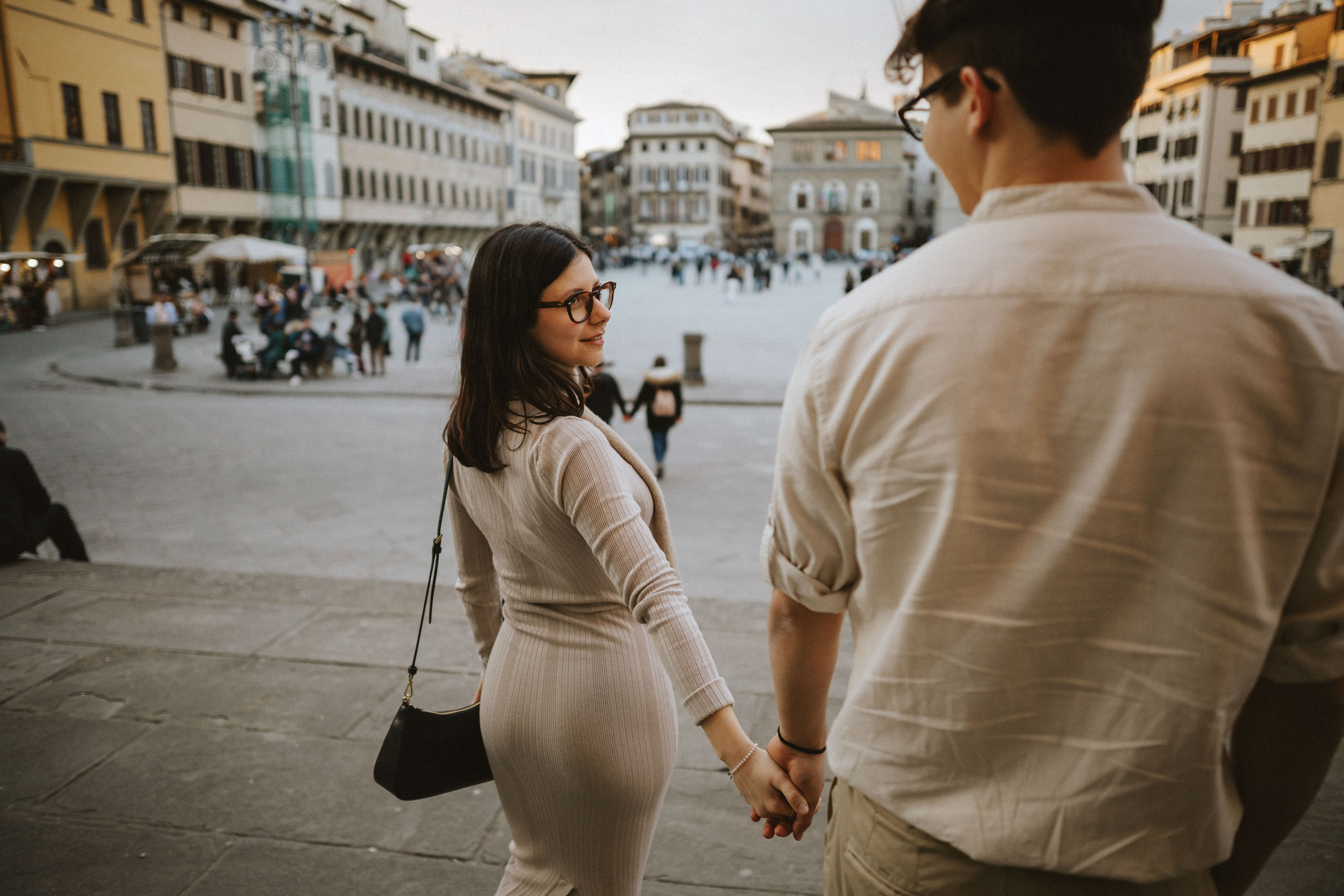 “Una coppia che cammina mano nella mano in una strada panoramica della città, con la donna che si guarda alle spalle verso l'uomo, sorridendo. Sullo sfondo, edifici e una vivace piazza pubblica”.
