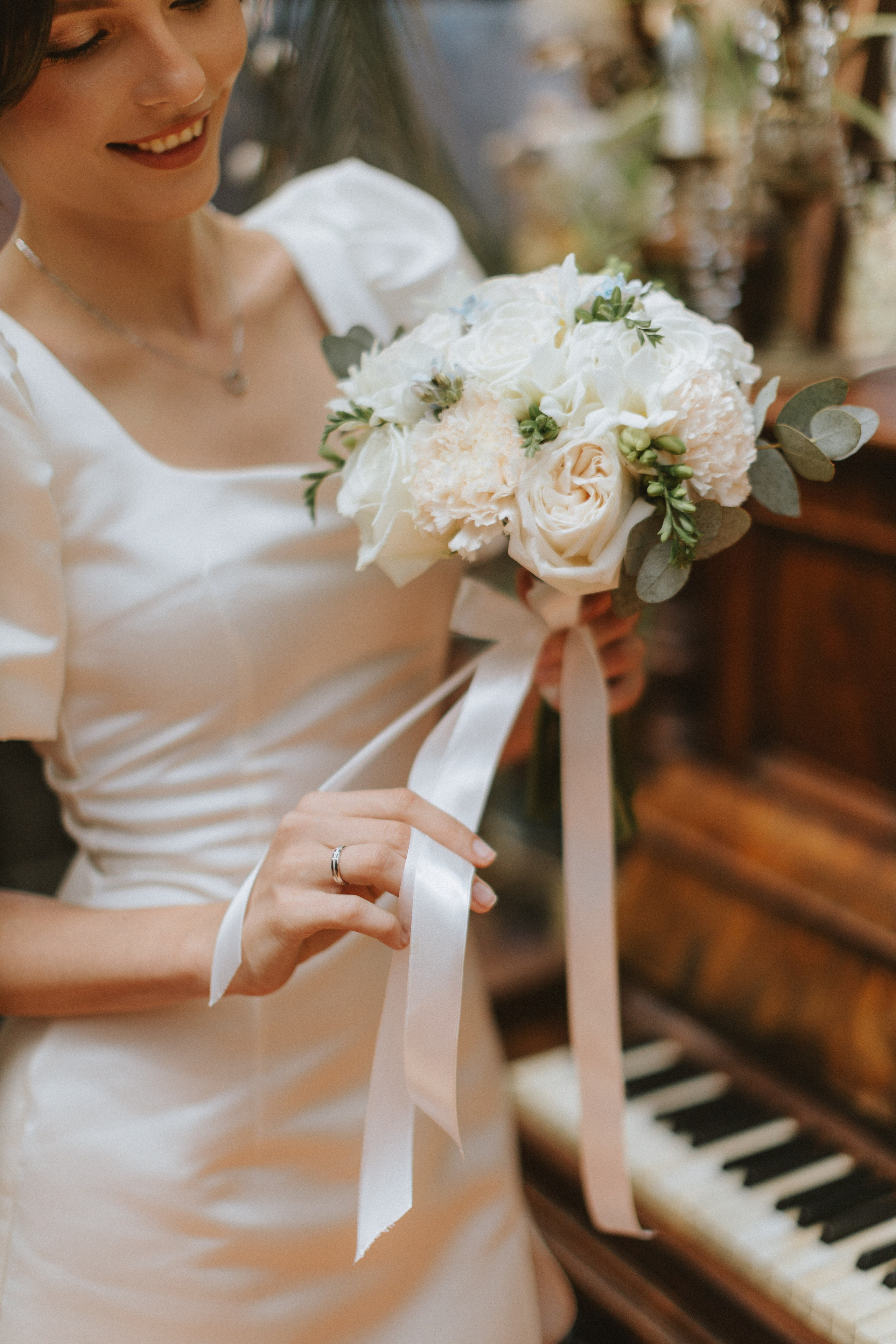 “La sposa aggiusta delicatamente i nastri del suo bouquet, sorridendo dolcemente mentre stringe i fiori bianchi e arrossati. Il pianoforte vintage sullo sfondo aggiunge un tocco classico, rendendo questo momento elegante prima della cerimonia nuziale”.