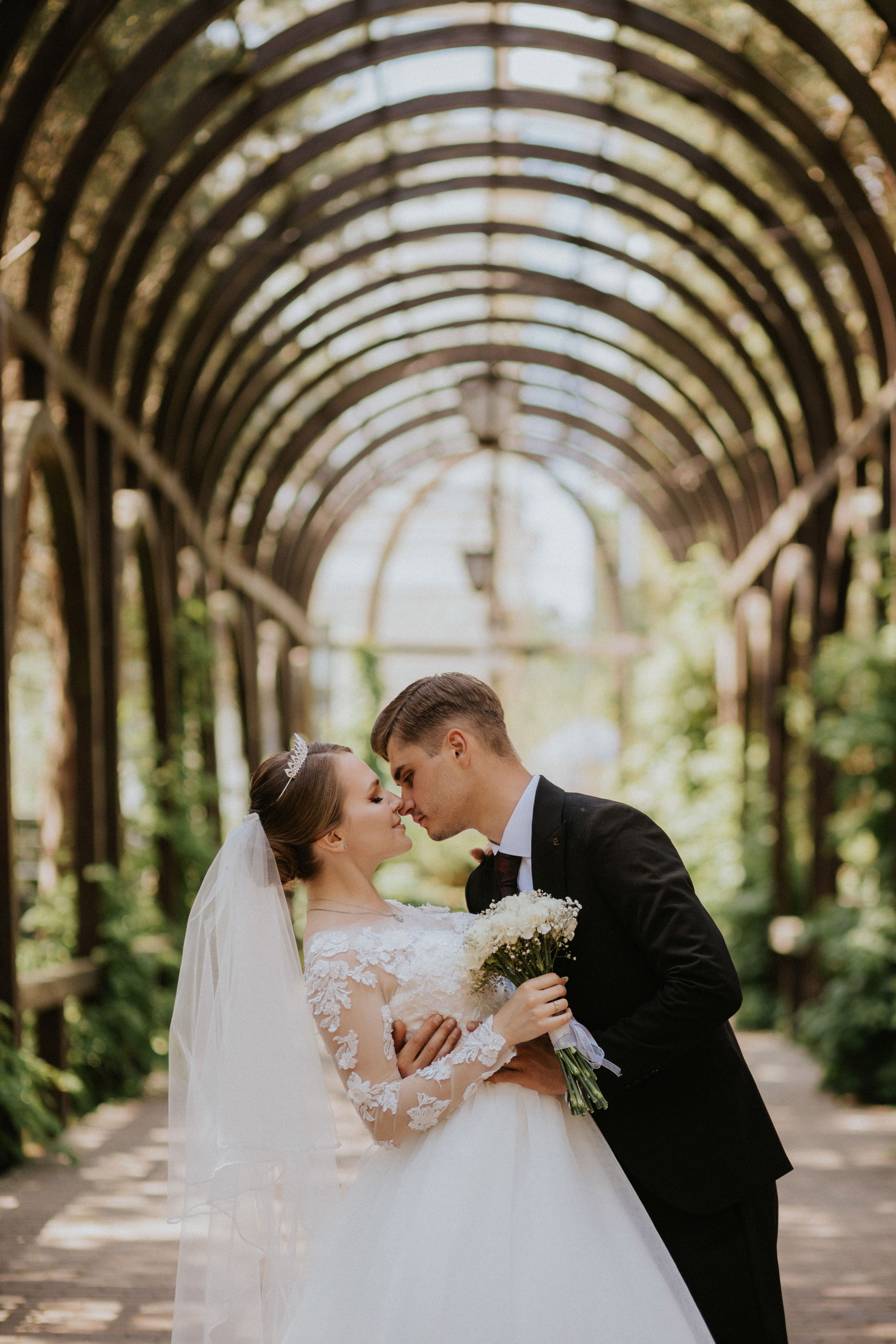 “Una coppia di sposi che condivide un momento di tenerezza sotto un viale ad arco, circondato da una vegetazione lussureggiante. La sposa, che indossa un delicato abito da sposa in pizzo con maniche lunghe e velo, tiene in mano un bouquet di fiori bianchi. Lo sposo, in un classico abito nero, le tocca delicatamente il viso mentre si sorridono a vicenda”.