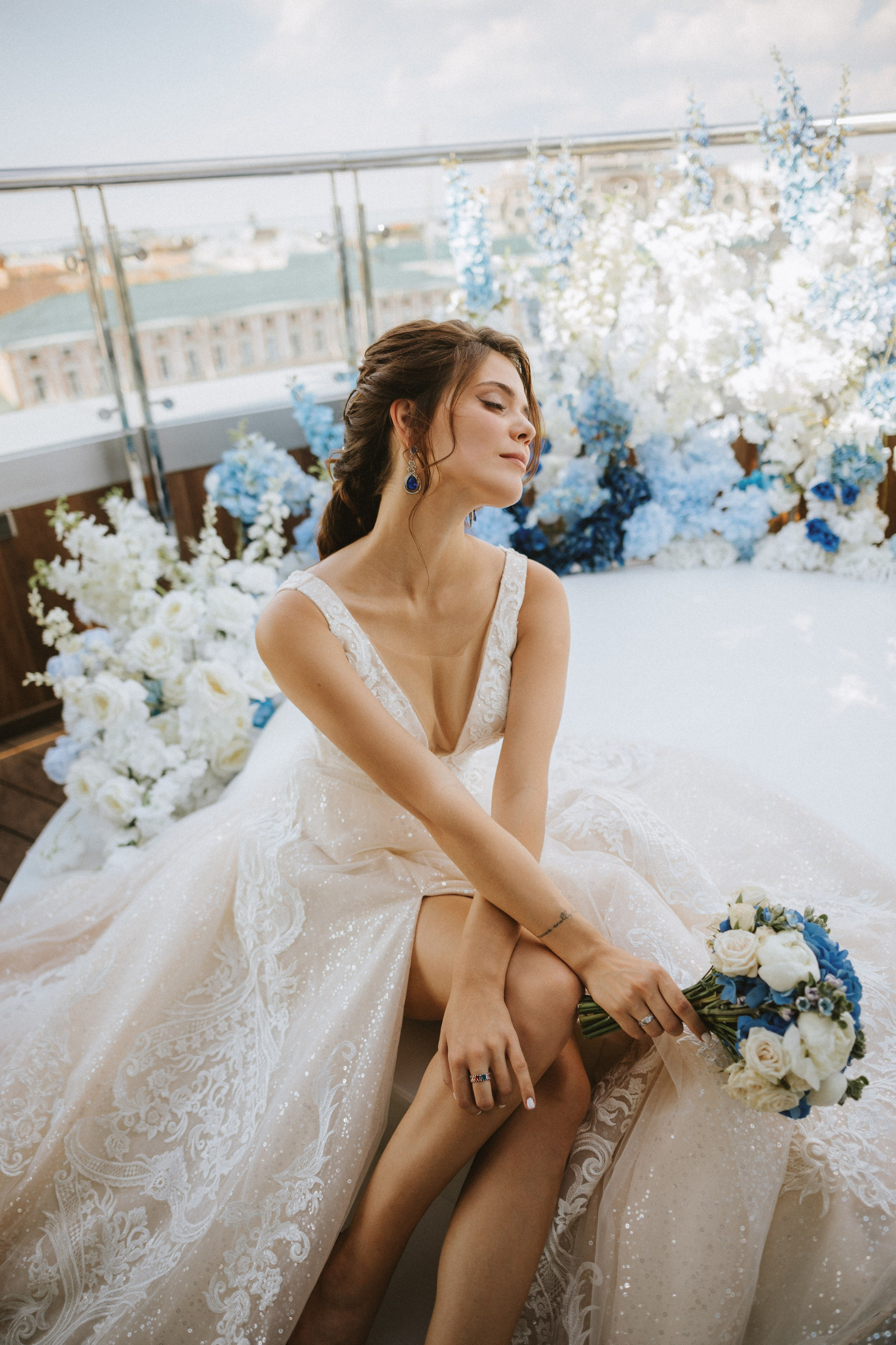 “La sposa, vestita con un abito scintillante, posa con grazia su uno sfondo di fiori bianchi e blu. Tiene in mano un bouquet di rose mentre guarda pensierosa in lontananza, trasudando eleganza e calma, perfetta per un elegante matrimonio sul tetto”.
