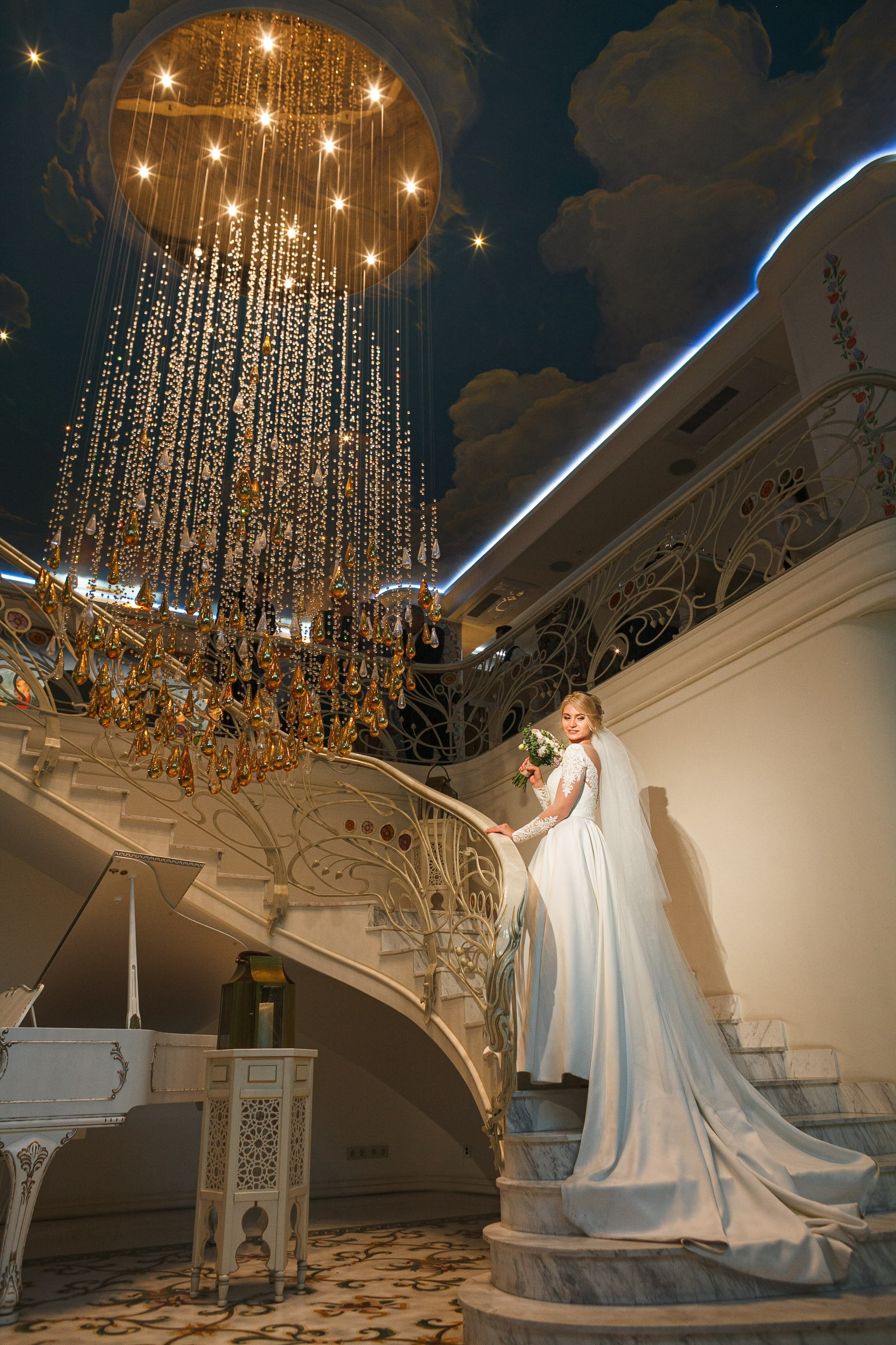 “Una sposa si erge con grazia su una scala, guardando la macchina fotografica mentre tiene in mano un bouquet di fiori. Indossa un elegante abito bianco con maniche lunghe in pizzo e il velo le ricade dietro. Sopra di lei, uno splendido lampadario con gocce d'oro a cascata illumina lo spazio e l'intricata ringhiera in ferro battuto della scala aggiunge un tocco di lusso. La stanza è decorata con ricchi dettagli, tra cui un soffitto dipinto con nuvole, che creano un'atmosfera regale e sofisticata”.