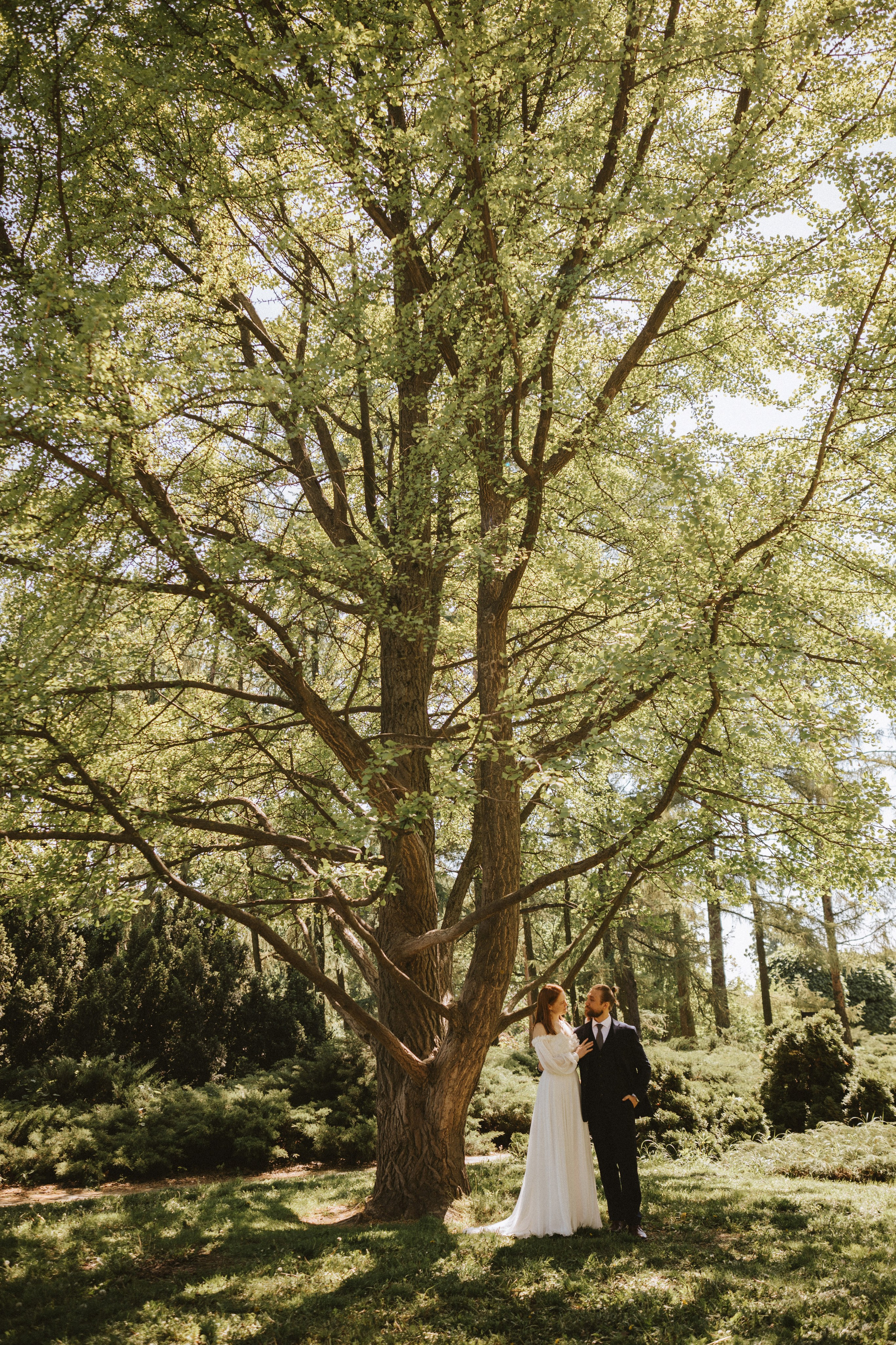 “Una sposa e uno sposo in piedi sotto un alto albero con foglie di un verde intenso. La sposa indossa un lungo e fluente abito bianco con maniche lunghe, mentre lo sposo è vestito con un abito nero con papillon. Si guardano amorevolmente, circondati da una vegetazione lussureggiante sullo sfondo”.