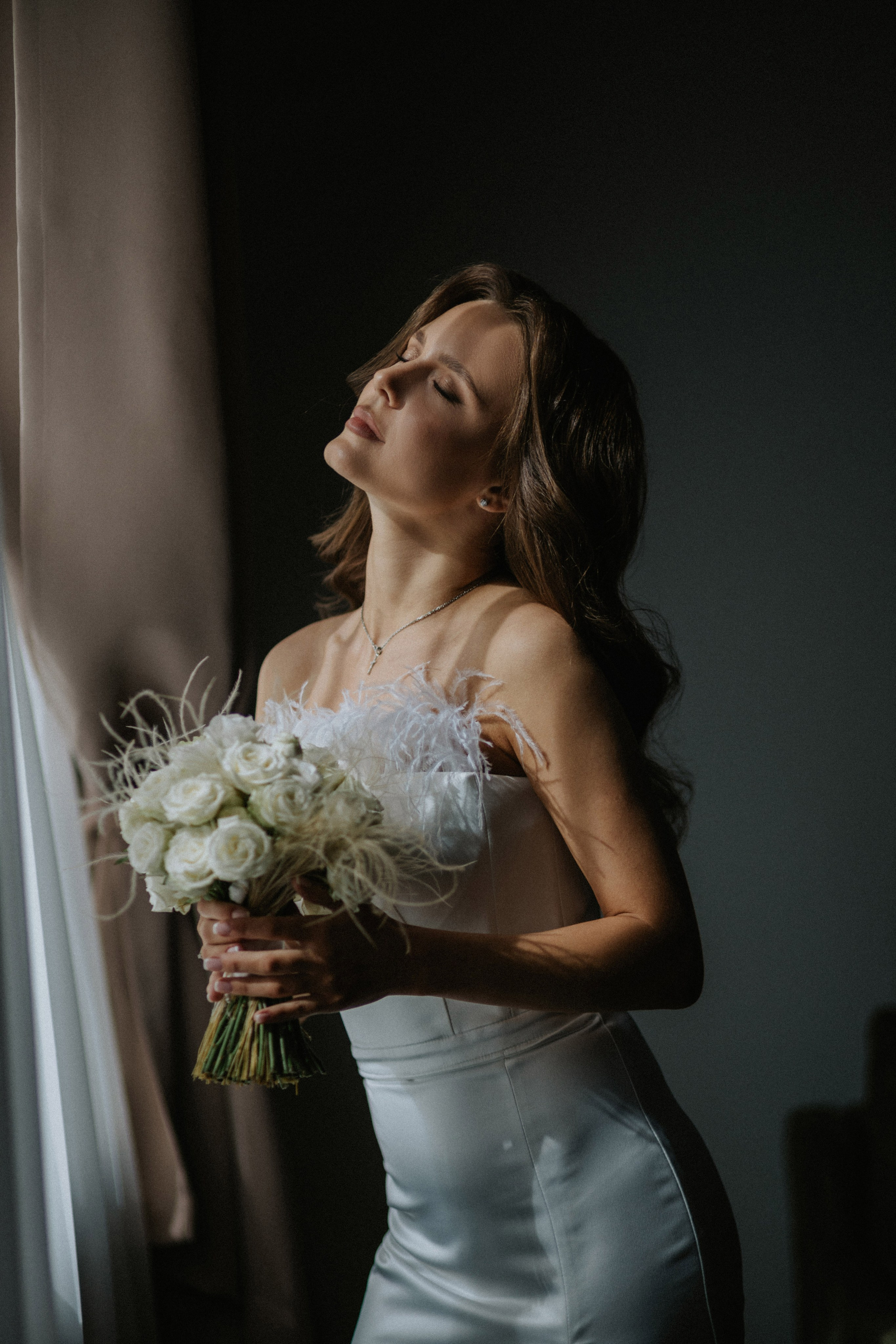 “Una sposa in un elegante abito bianco con accenti di piume chiude gli occhi, assaporando il momento. Tiene delicatamente tra le mani un bouquet di rose bianche, mentre una luce naturale soffusa le illumina il viso”.