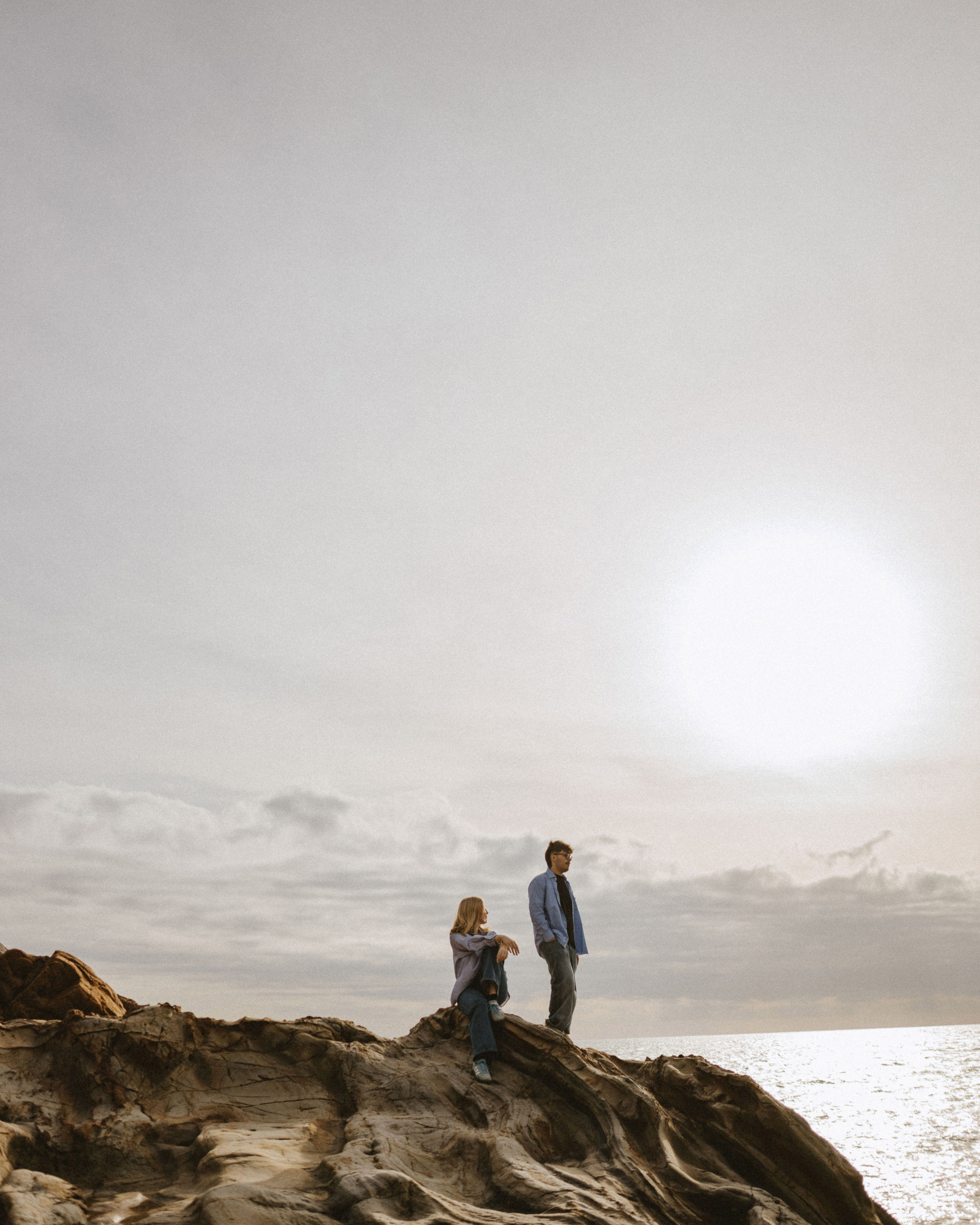 “The couple sitting and standing on the rocks by the shore, enjoying the view of the ocean under the bright sunlight.”