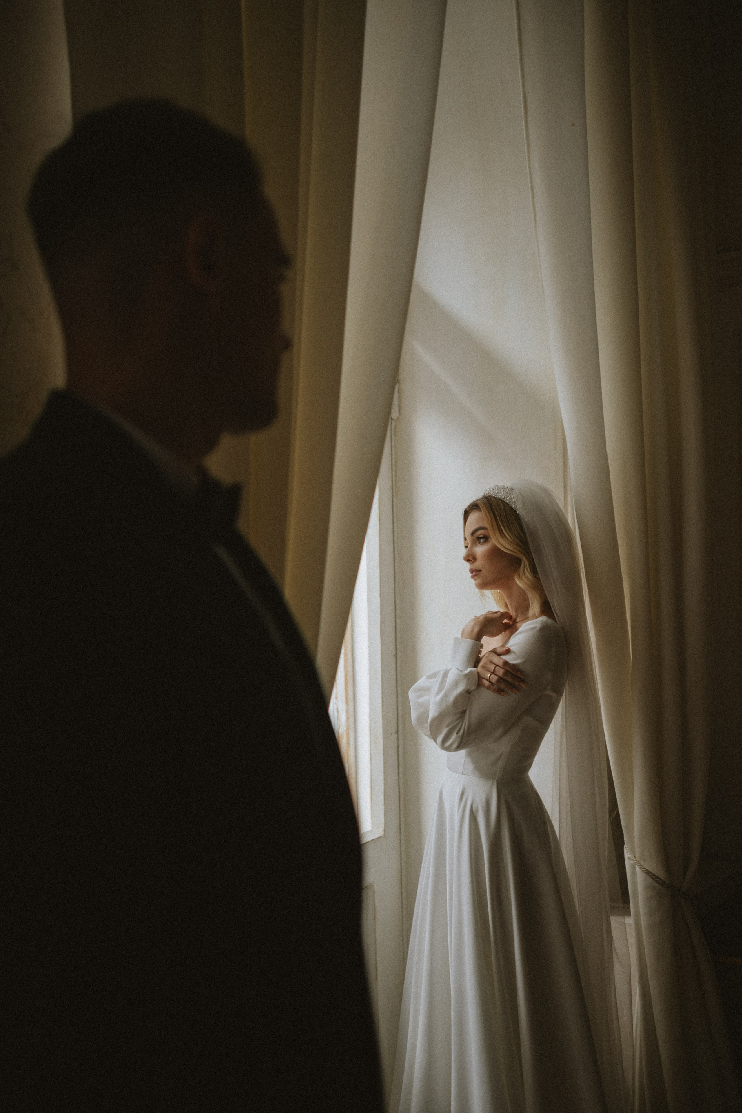 “Fotografo di matrimonio che cattura un momento intimo della sposa nel suo elegante abito da sposa bianco e velo, in piedi accanto a una finestra con una luce naturale morbida. La silhouette dello sposo si intravede sullo sfondo, aggiungendo un'atmosfera romantica. Un bellissimo scatto per un matrimonio in Toscana”.