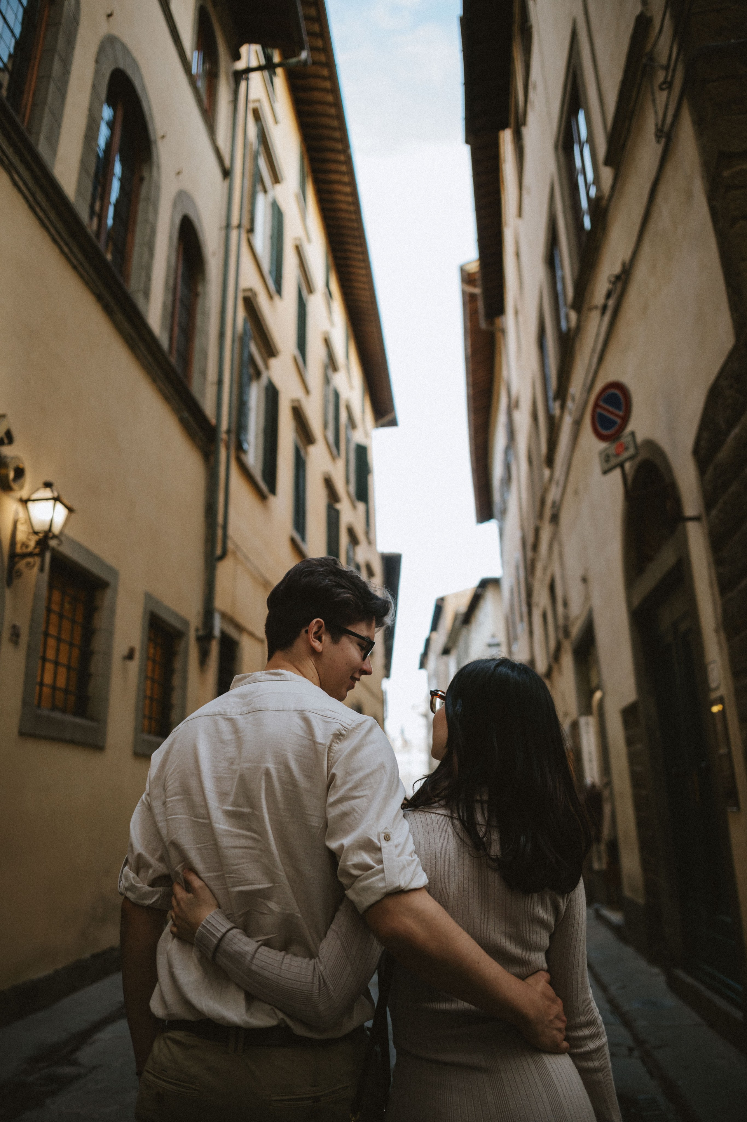“Una coppia che cammina in una strada stretta, tenendosi per mano con l'uomo che abbraccia la donna da dietro, entrambi si guardano”.