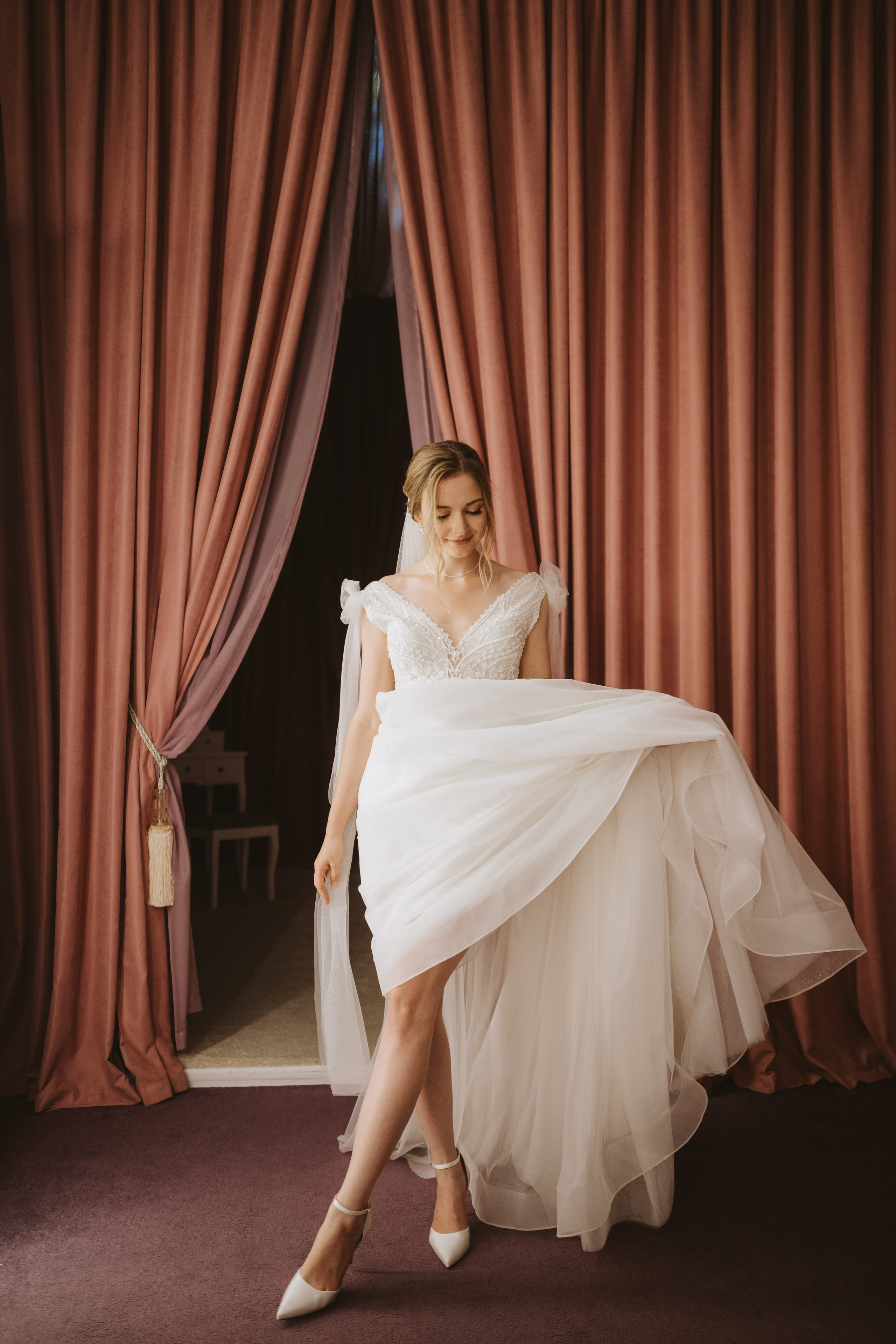 “Una sposa in abito bianco con delicati dettagli in pizzo solleva la gonna del suo abito, sorridendo dolcemente mentre si trova davanti a tende di velluto rosa. Indossa un velo e tacchi bianchi, emanando eleganza nella stanza illuminata da tinte pastello”.