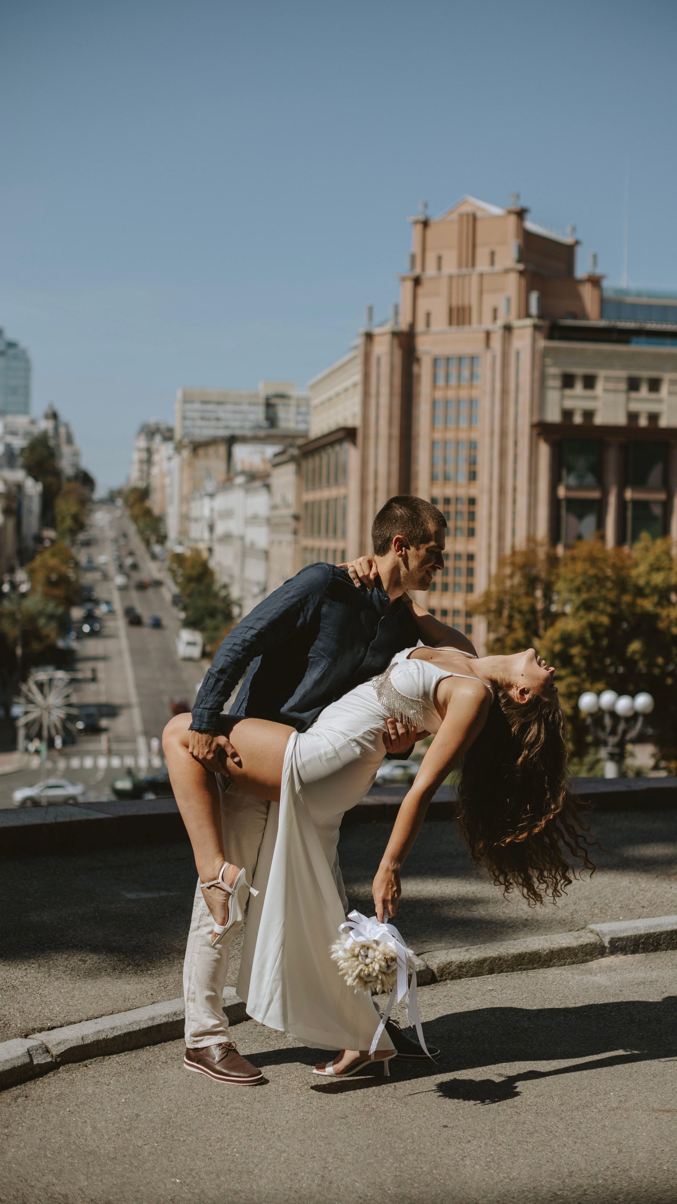 “Una gioiosa coppia di sposi in un contesto urbano. Lo sposo tiene la sposa in un tuffo giocoso, con la testa inclinata all'indietro mentre ride. La sposa indossa un elegante abito bianco con un dettaglio di perline, mentre lo sposo è vestito in modo casual con una camicia marina e pantaloni leggeri. Lo sfondo è rappresentato da una vivace strada cittadina con alti edifici e un cielo limpido e azzurro”.