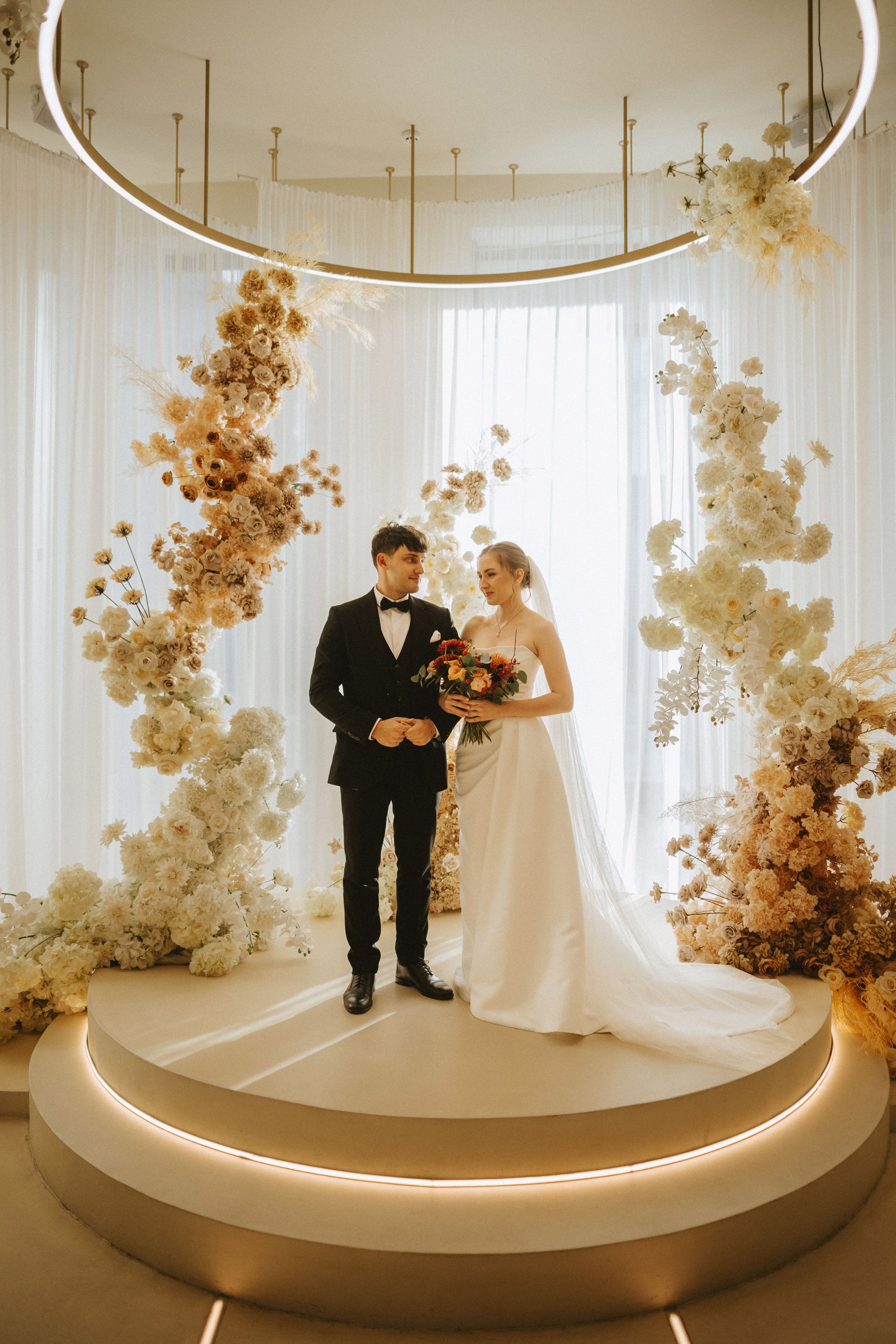 “Una sposa e uno sposo si trovano insieme su una piattaforma rialzata in un luogo splendidamente decorato. La sposa indossa un semplice ed elegante abito bianco con velo e tiene in mano un bouquet di fiori colorati. Lo sposo, vestito con uno smoking nero con papillon, le sorride. Sono circondati da composizioni floreali nei toni tenui del bianco, del crema e del beige, con un moderno lampadario circolare in alto”.