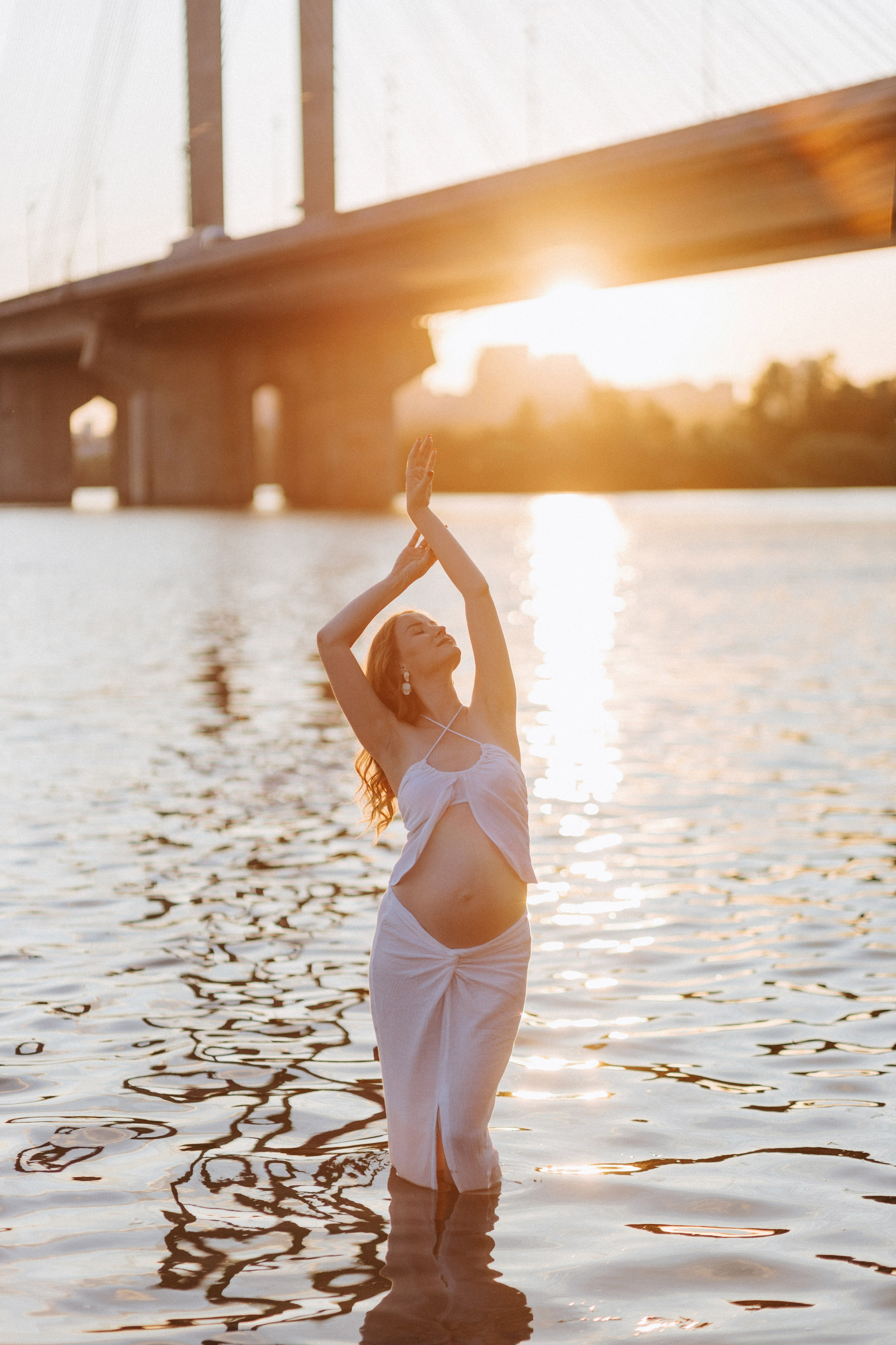 “Una donna incinta in abito bianco in piedi nell'acqua con le braccia sollevate con grazia, stagliata contro il sole al tramonto e un ponte sullo sfondo”.