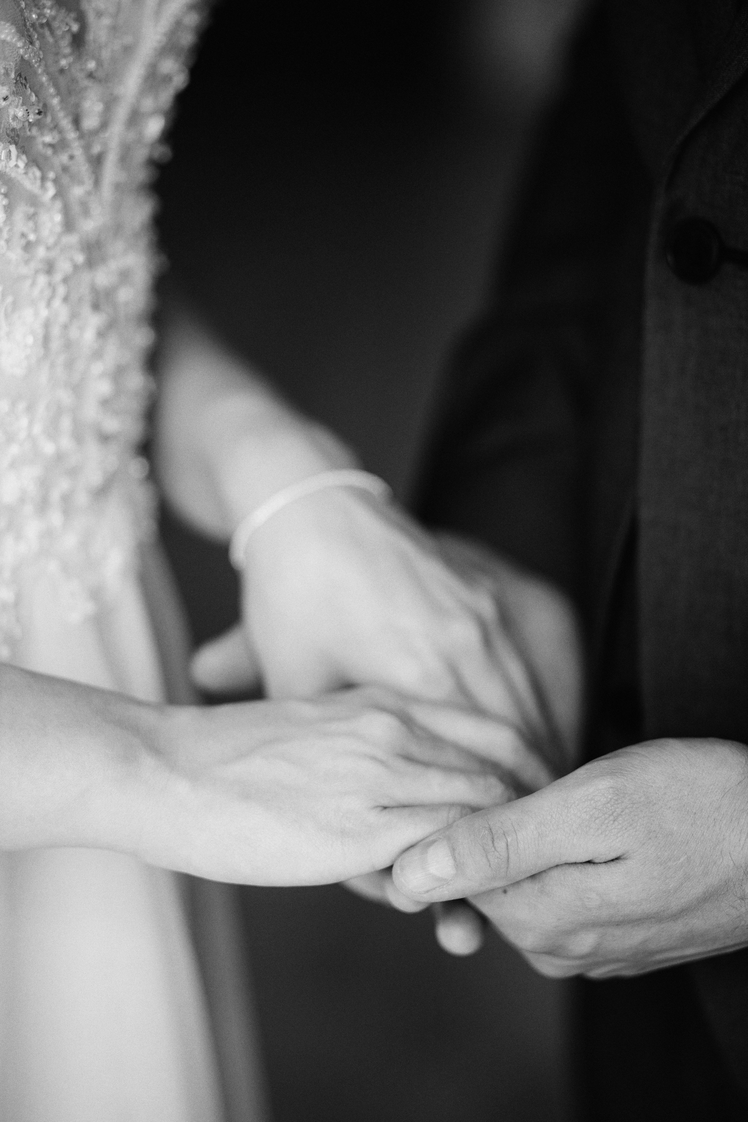 “Un primo piano in bianco e nero delle mani di una coppia, che si stringono dolcemente. Una mano è ornata da una fede nuziale, simbolo del loro legame, mentre sullo sfondo si intravede la morbida consistenza dell'abito della sposa.”