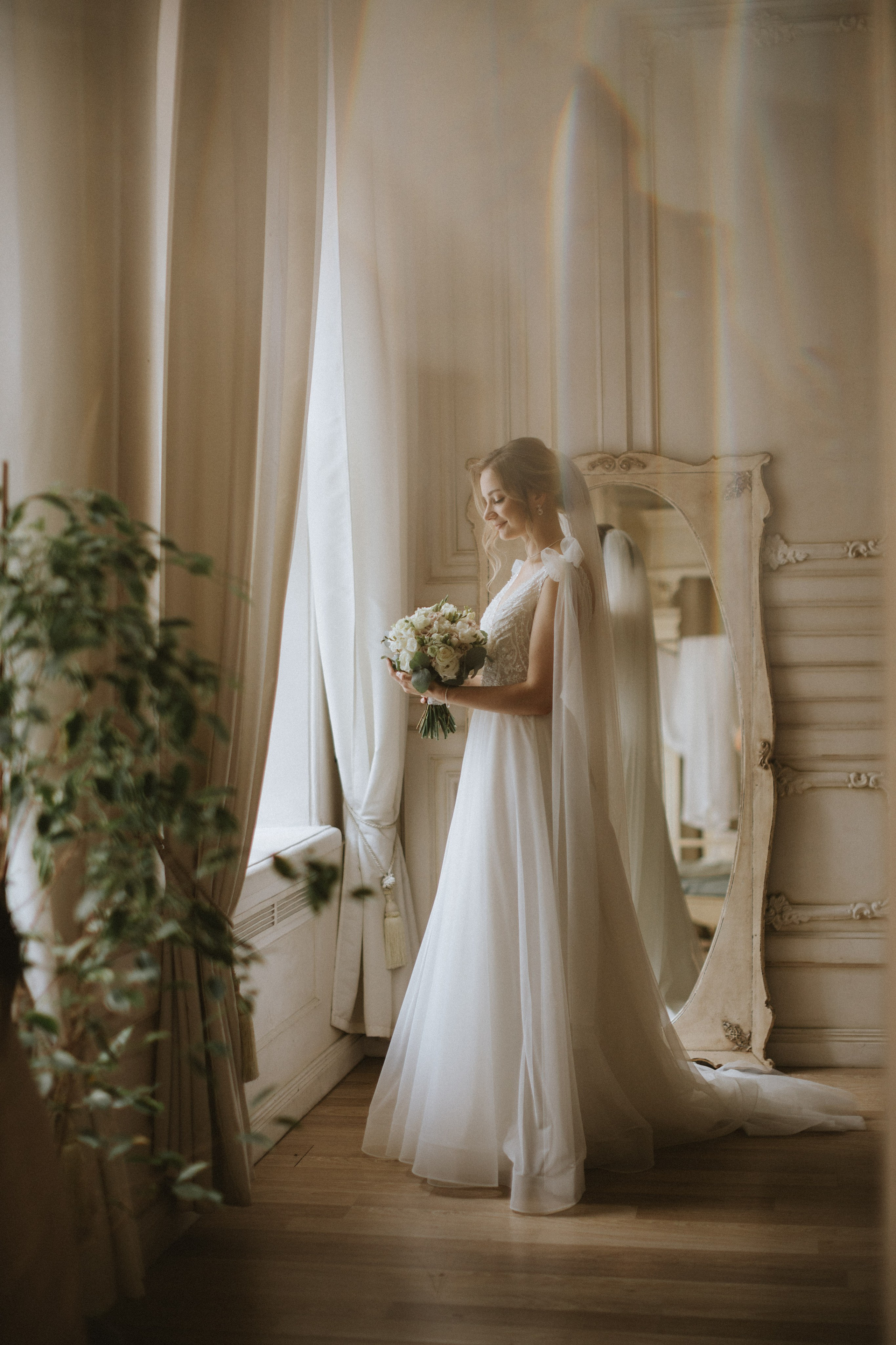 “Una sposa in un abito morbido e fluente sta davanti a un'ampia finestra, guardando il suo bouquet con un sorriso sereno. Il velo le scende delicatamente sulla schiena e l'abito scorre elegantemente sul pavimento. La luce soffusa che filtra dalle tende crea un'atmosfera romantica, mentre il riflesso nello specchio vicino aggiunge un tocco di grazia al momento. La scena è tranquilla e piena di calore”.