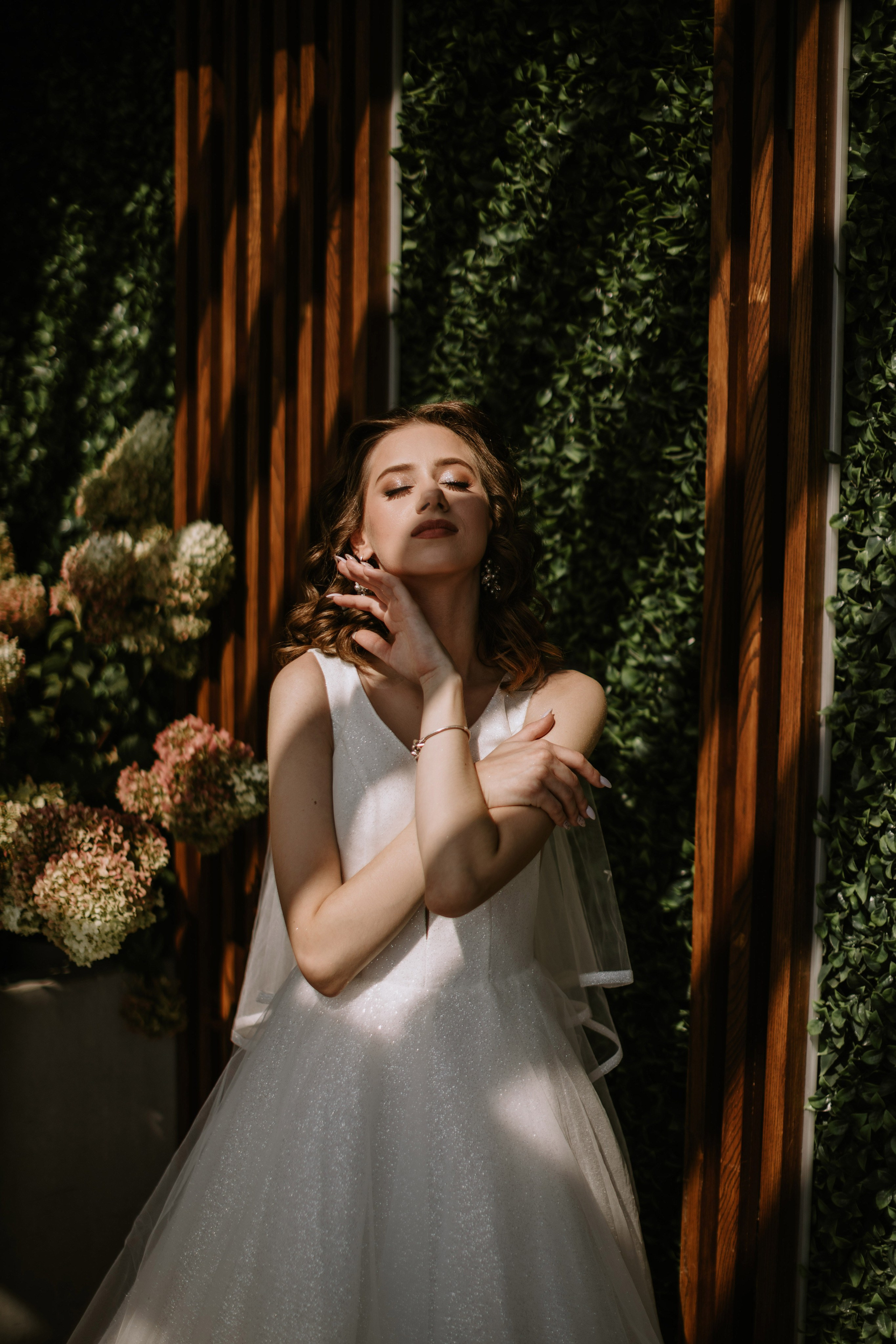 “Una sposa in un abito bianco scintillante si erge con grazia davanti a uno sfondo di verde lussureggiante. La luce soffusa del sole crea un bellissimo gioco di ombre sul suo viso e sull'abito mentre incrocia delicatamente le braccia, irradiando un'aura serena ed elegante.”