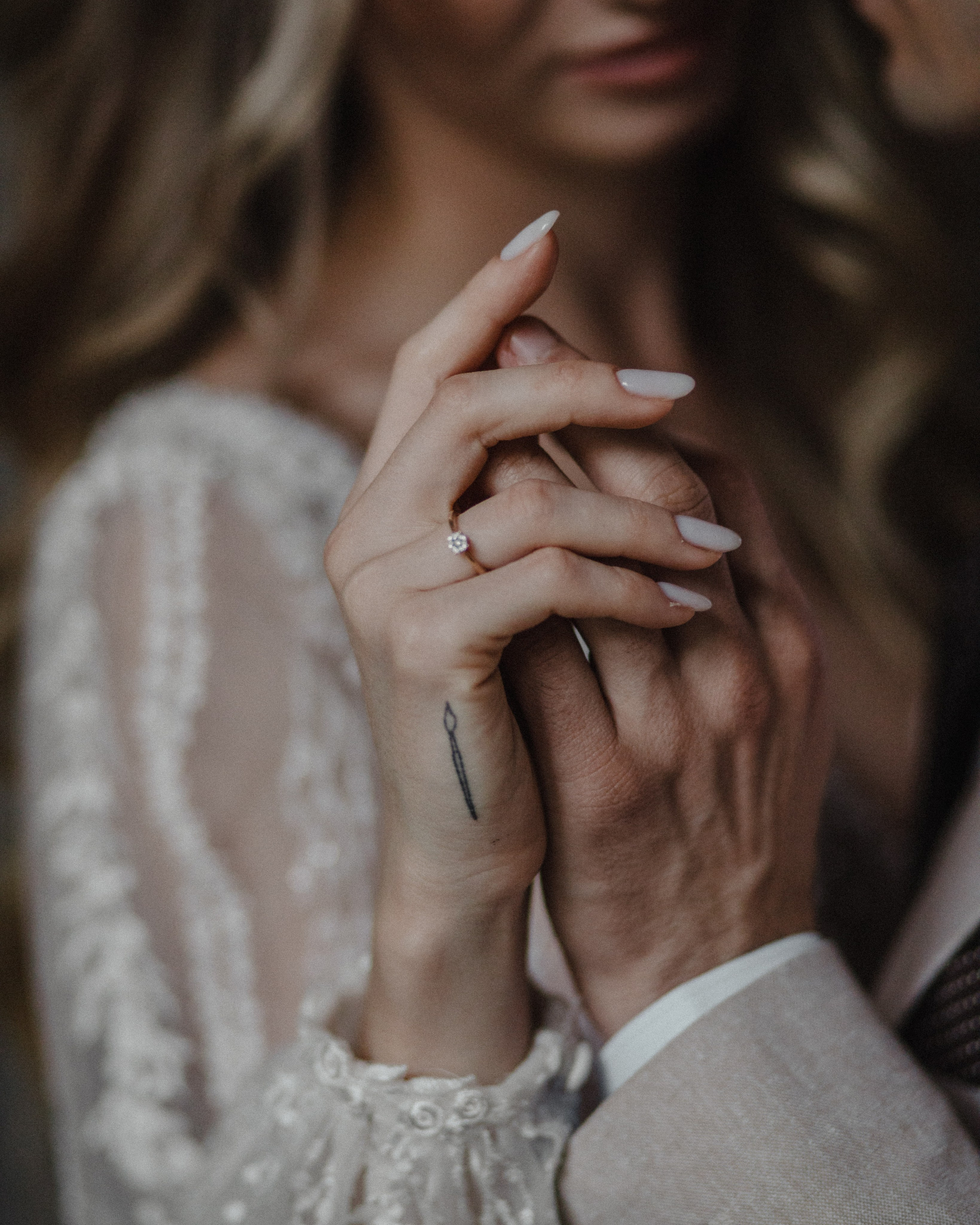 “Primo piano di una coppia che si tiene per mano, mostrando l'anello di fidanzamento della sposa e un tatuaggio sulla sua mano. La coppia sta condividendo un momento di intimità, catturato durante un servizio fotografico di matrimonio”.