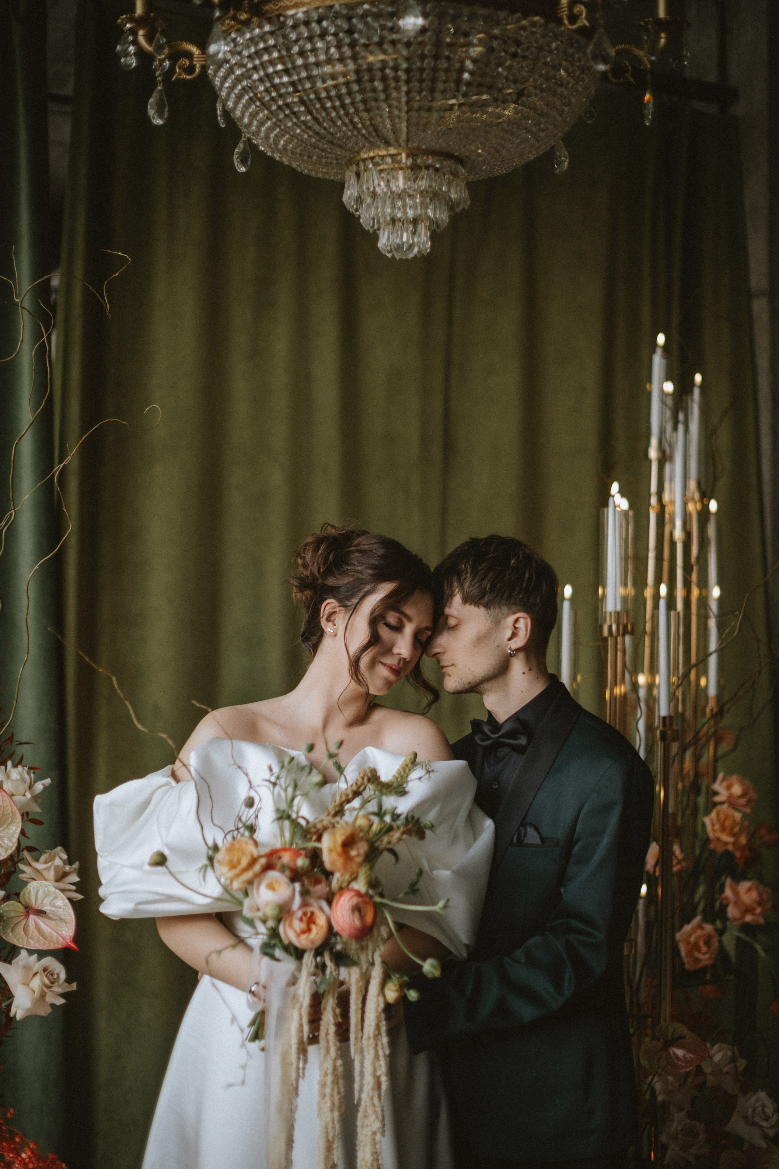 “Una sposa e uno sposo sono vicini in una posa intima, condividendo un momento di tenerezza. La sposa, vestita con uno splendido abito bianco off-shoulder, tiene in mano un bellissimo bouquet di fiori dai toni tenui. Lo sposo, in smoking verde scuro con papillon, appoggia delicatamente la fronte su quella di lei. I due si trovano sotto un grande lampadario, con luci soffuse che sottolineano il loro romantico abbraccio. Alti ed eleganti portacandele li circondano, aggiungendo un'atmosfera sofisticata”.