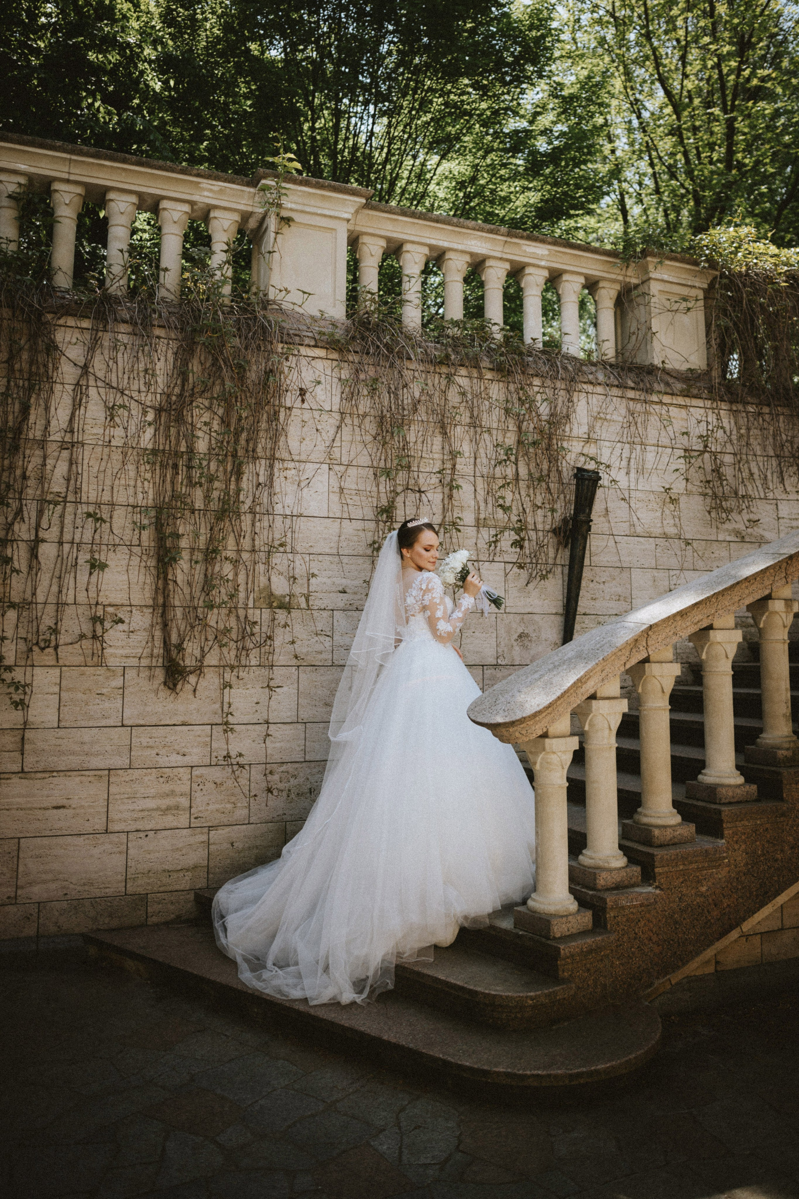 “Una bellissima sposa in un fluente abito bianco ornato da dettagli in pizzo si trova su una scalinata, con in mano un bouquet di fiori bianchi. La sposa guarda in basso con un sorriso sereno, il suo velo scorre dietro di lei mentre si trova contro un muro di pietra strutturato coperto di edera. La scala e il verde circostante creano un'atmosfera tranquilla e romantica”.