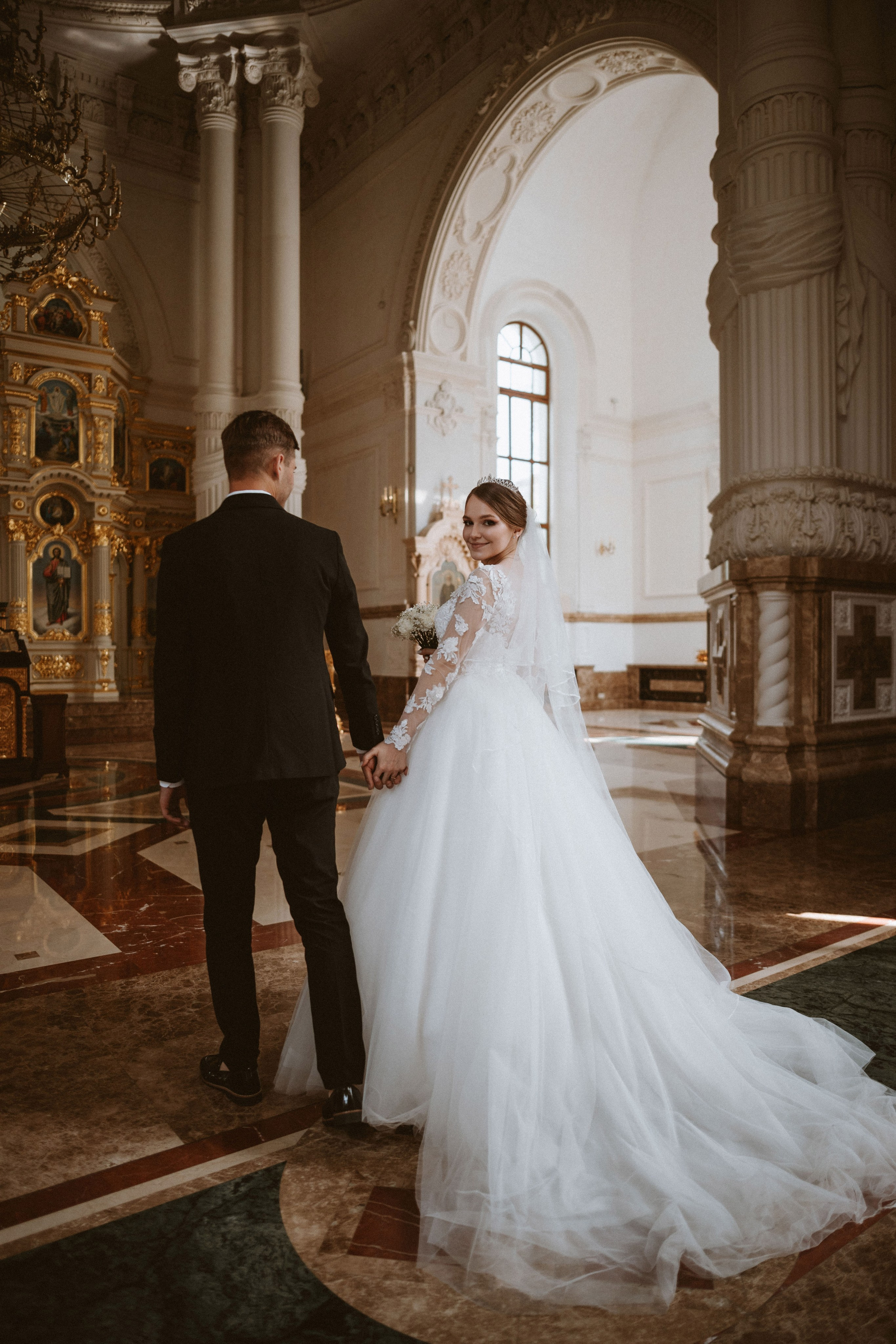 “Una sposa e uno sposo che si tengono per mano, camminando in una bellissima e grandiosa chiesa. La sposa, che indossa un abito di pizzo a maniche lunghe con una gonna voluminosa, sorride alla telecamera da sopra la spalla. Lo sposo, vestito con un classico abito nero, cammina accanto a lei, con uno sguardo alle decorazioni dorate della chiesa e alla luce che filtra dalle finestre alle loro spalle”.