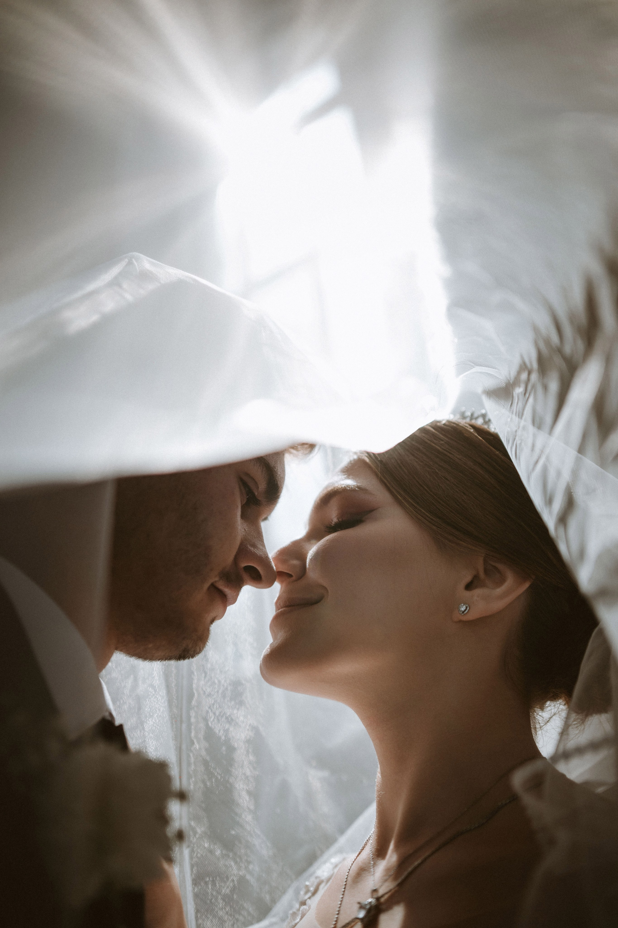 “Una sposa e uno sposo che condividono un momento di tenerezza, sorridendosi l'un l'altro mentre sono in piedi sotto un velo. La luce morbida proveniente da dietro crea un bagliore romantico ed etereo, catturando l'intimità del momento”.