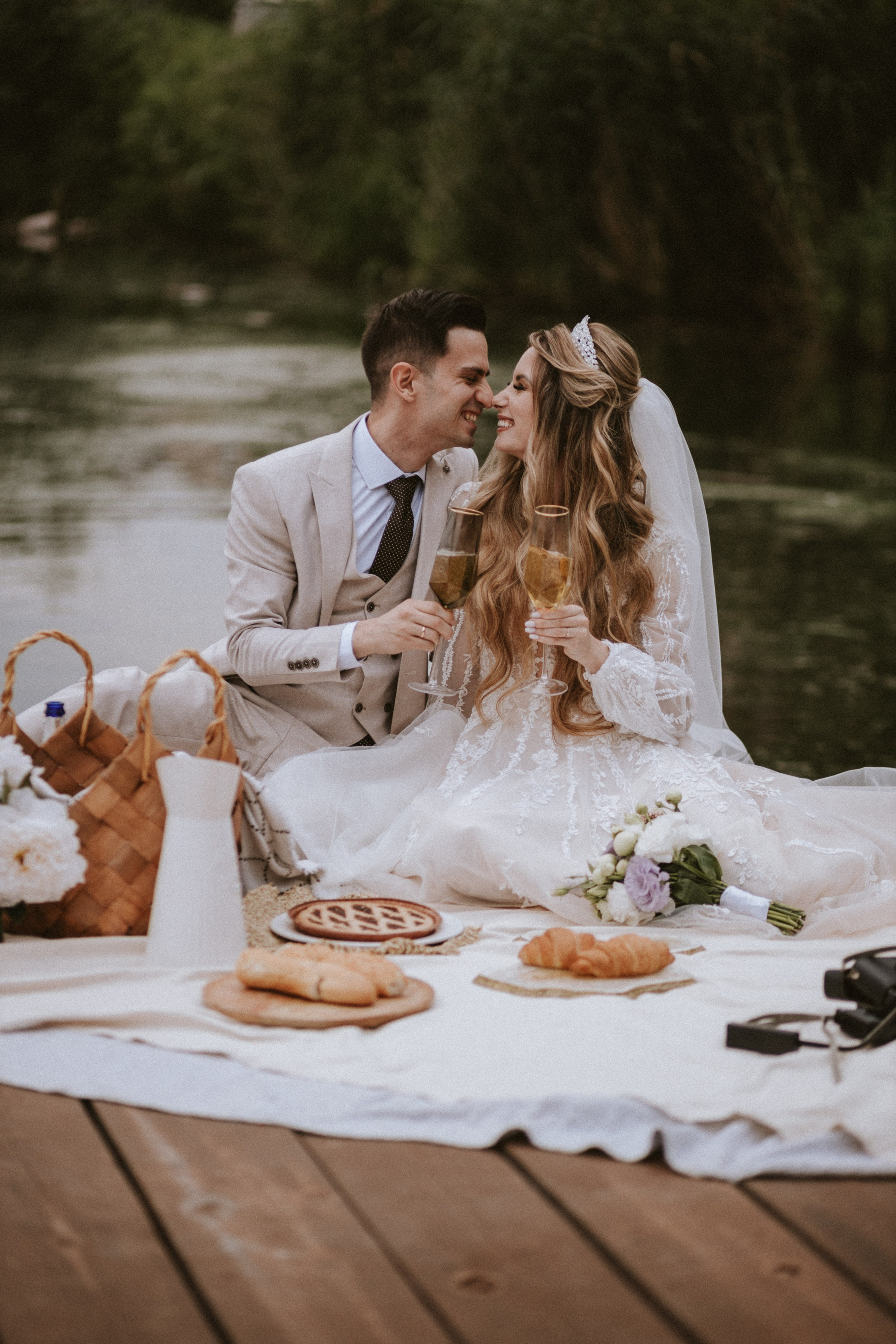 Stylish wedding couple enjoying picnic with sparkling wine and croissants on the pier