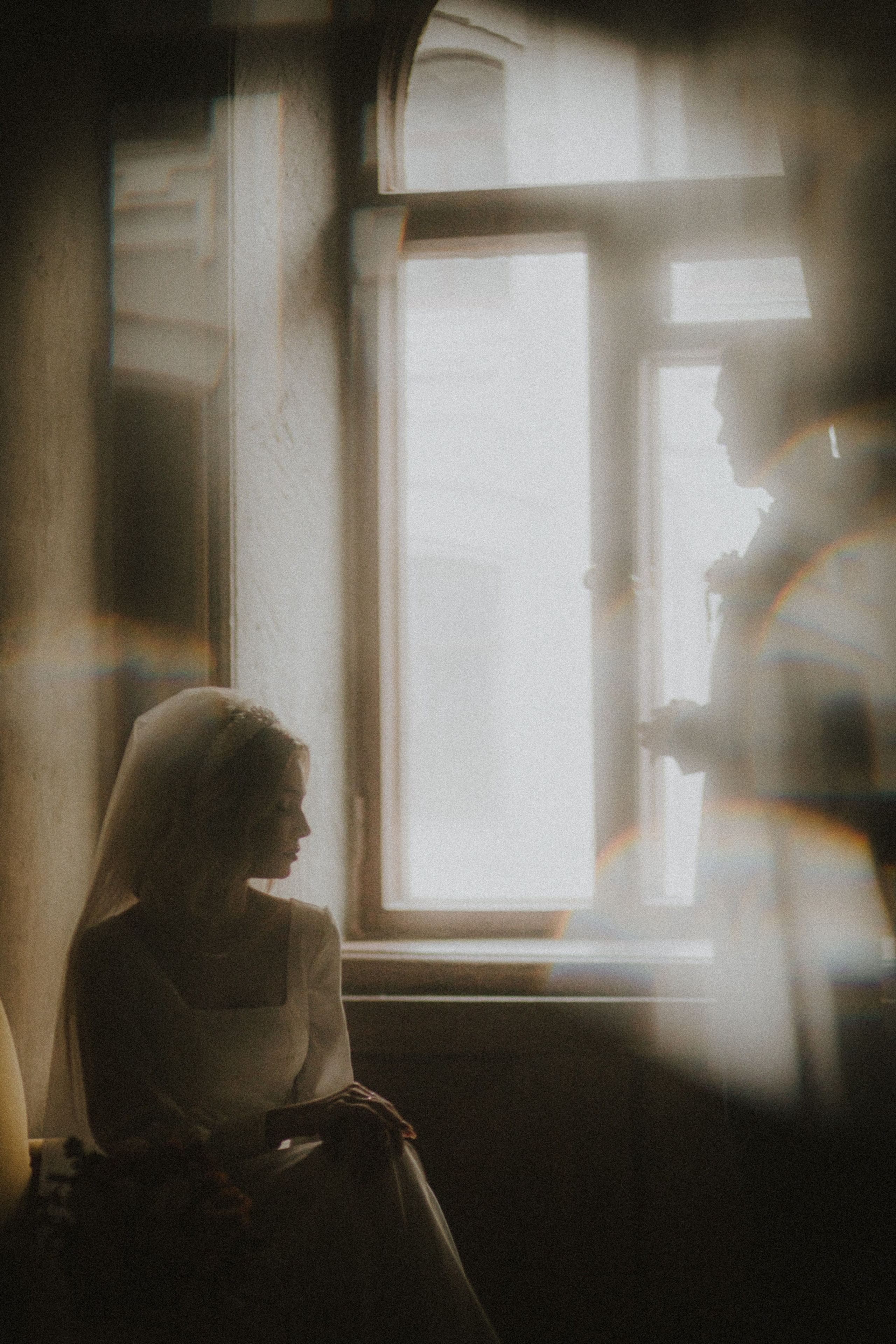 “Una sposa seduta vicino a una finestra, immersa in una luce soffusa, con un'espressione sognante e serena. La sagoma del suo compagno è visibile sullo sfondo, morbidamente sfocata, mentre la luce del sole proietta un delicato bagliore intorno a loro”.