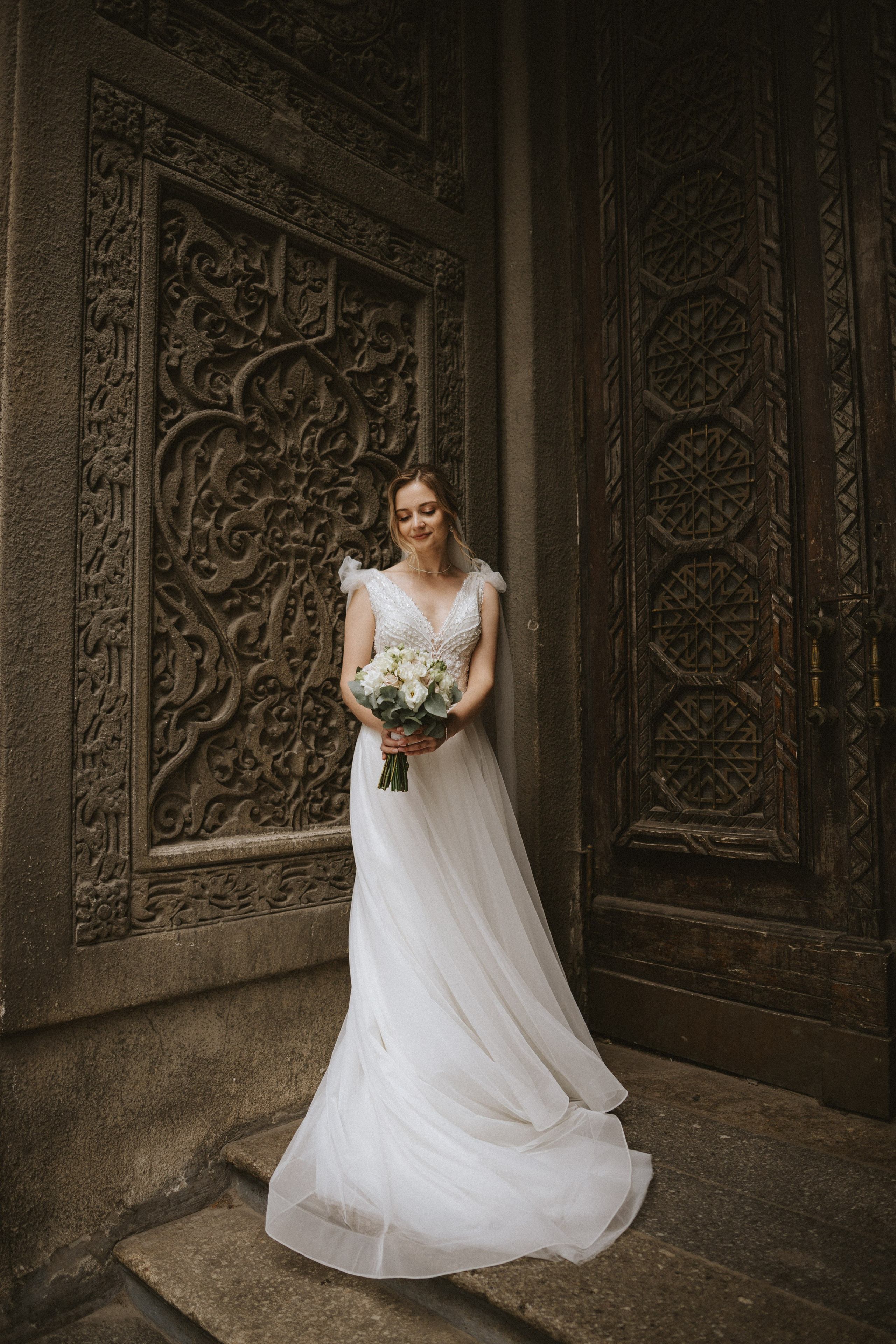 “La sposa si erge con grazia contro una porta di legno ornata, tenendo in mano un bouquet di fiori bianchi e verdi. Il suo elegante abito da sposa le scorre magnificamente intorno, mentre gli intricati dettagli della porta fanno da splendida cornice a questo momento senza tempo”.