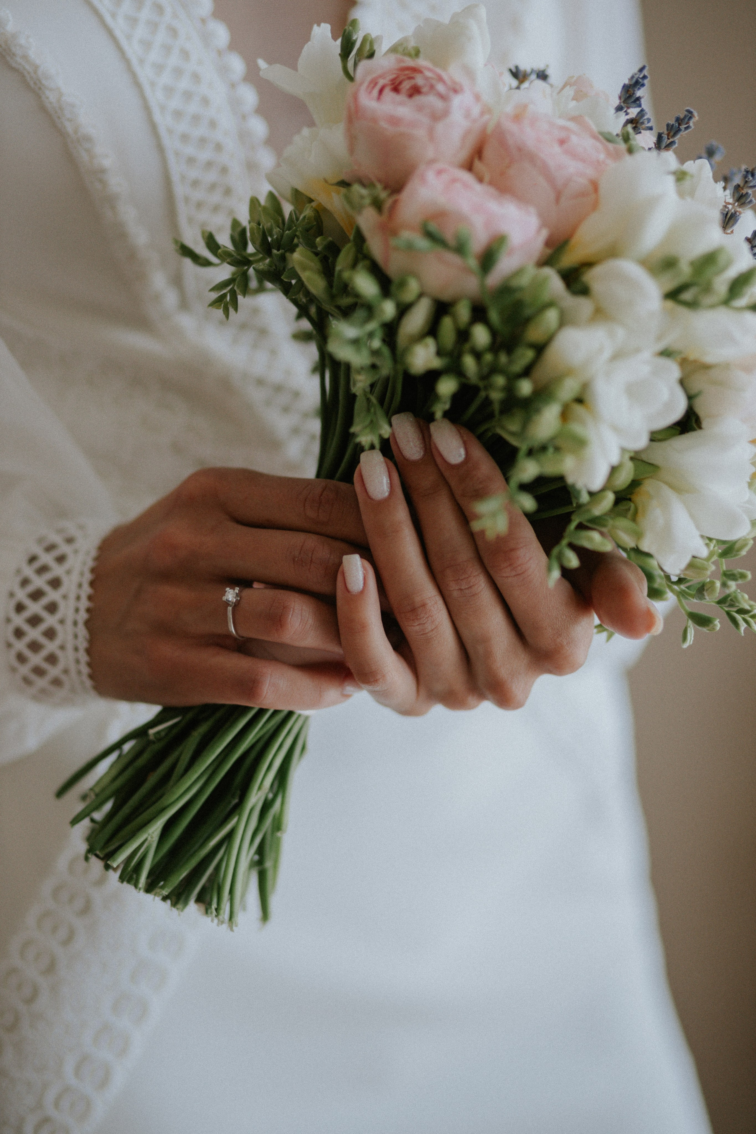 “Primo piano delle mani di una sposa che reggono un bouquet di delicate rose rosa, fiori bianchi e lavanda, con un'attenzione particolare all'anello di fidanzamento. Le unghie ben curate della sposa sono dipinte con toni morbidi e neutri”.