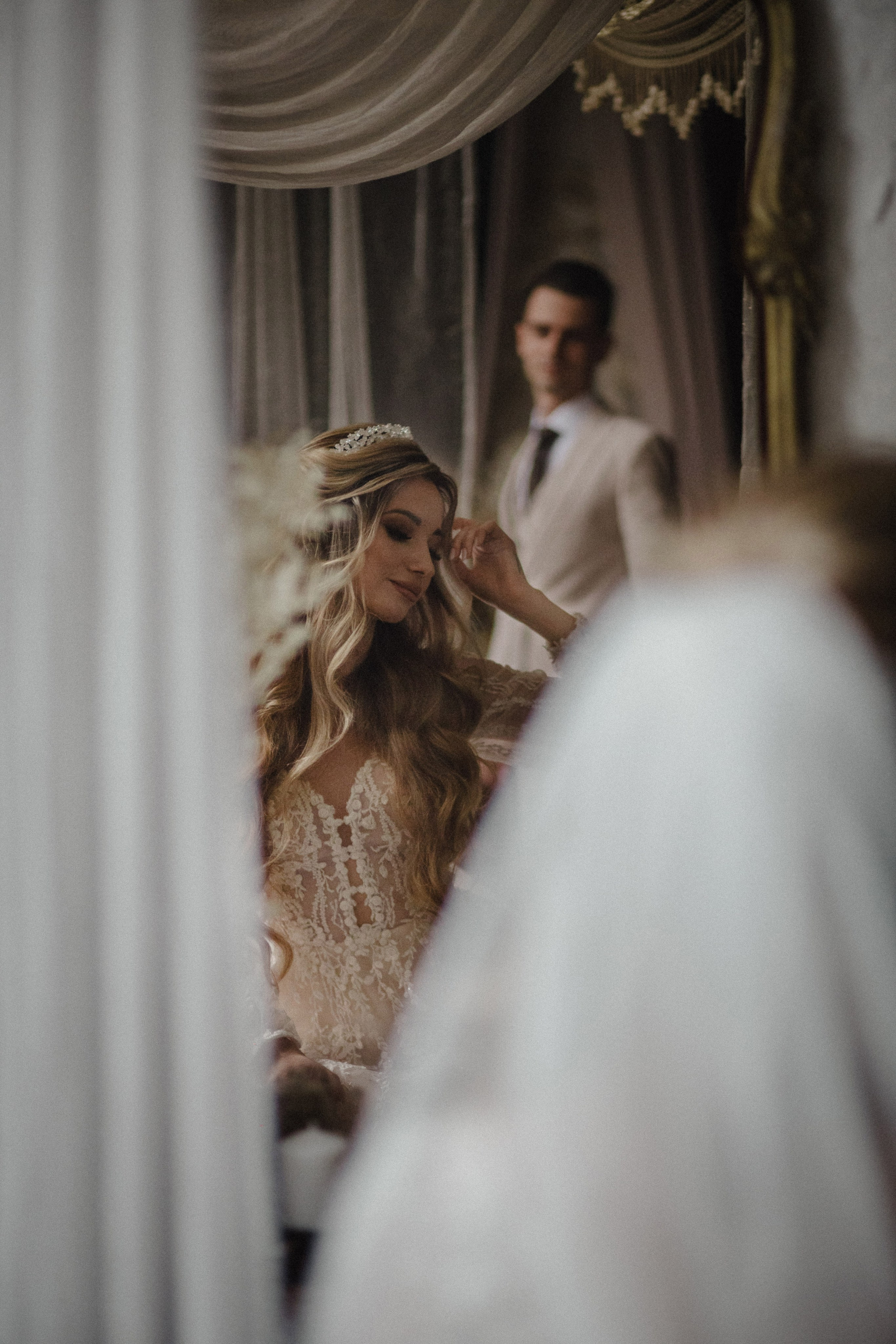 “Una sposa in un delicato abito di pizzo con diadema appare serena e pensierosa mentre si aggiusta i capelli. Il riflesso dello sposo si intravede sullo sfondo, aggiungendo un'atmosfera romantica al momento”.