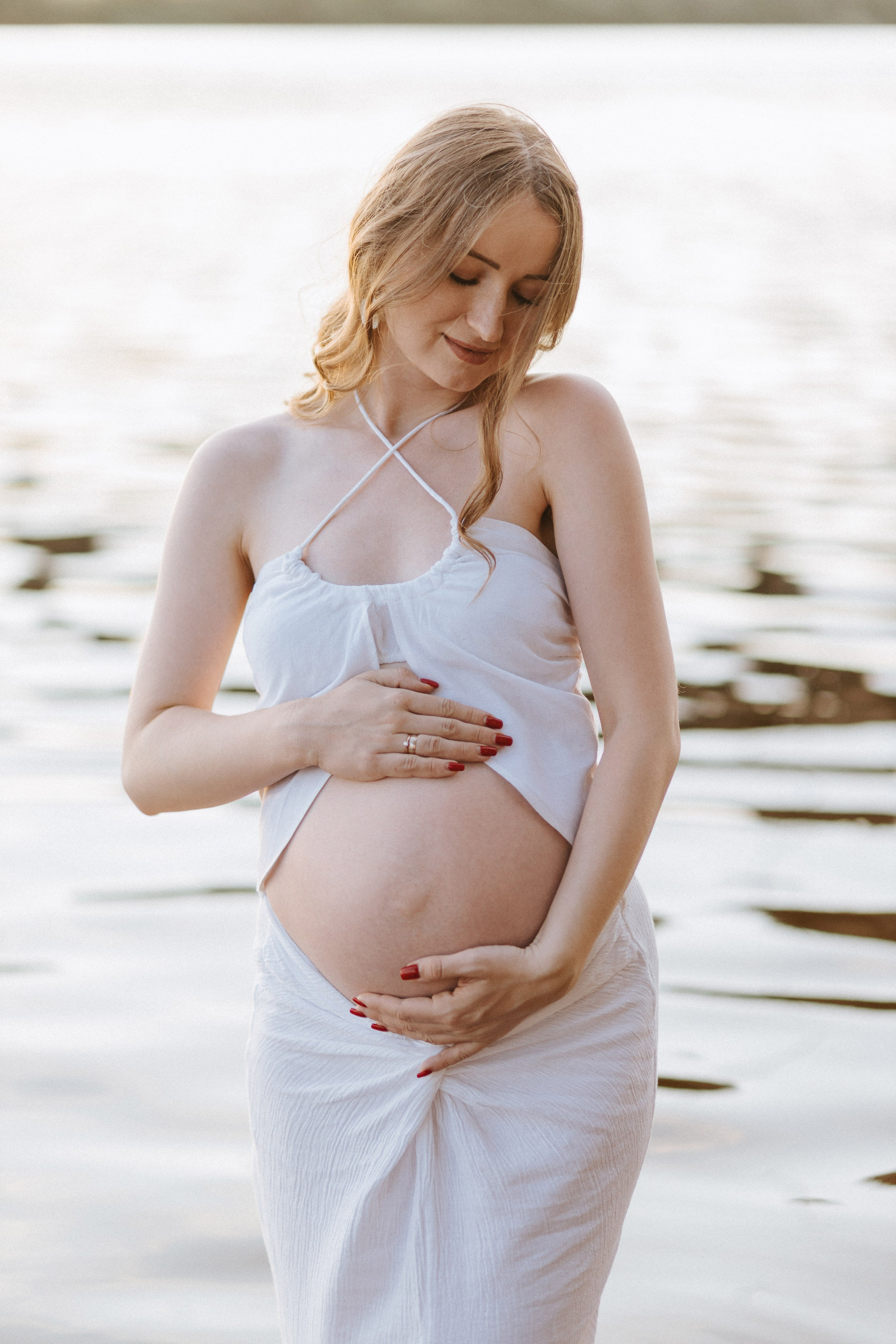 “Una donna incinta in piedi vicino all'acqua, che si tiene delicatamente la pancia indossando un abito bianco. Ha un'espressione serena, con gli occhi chiusi e i capelli che fluiscono dolcemente nella brezza”.