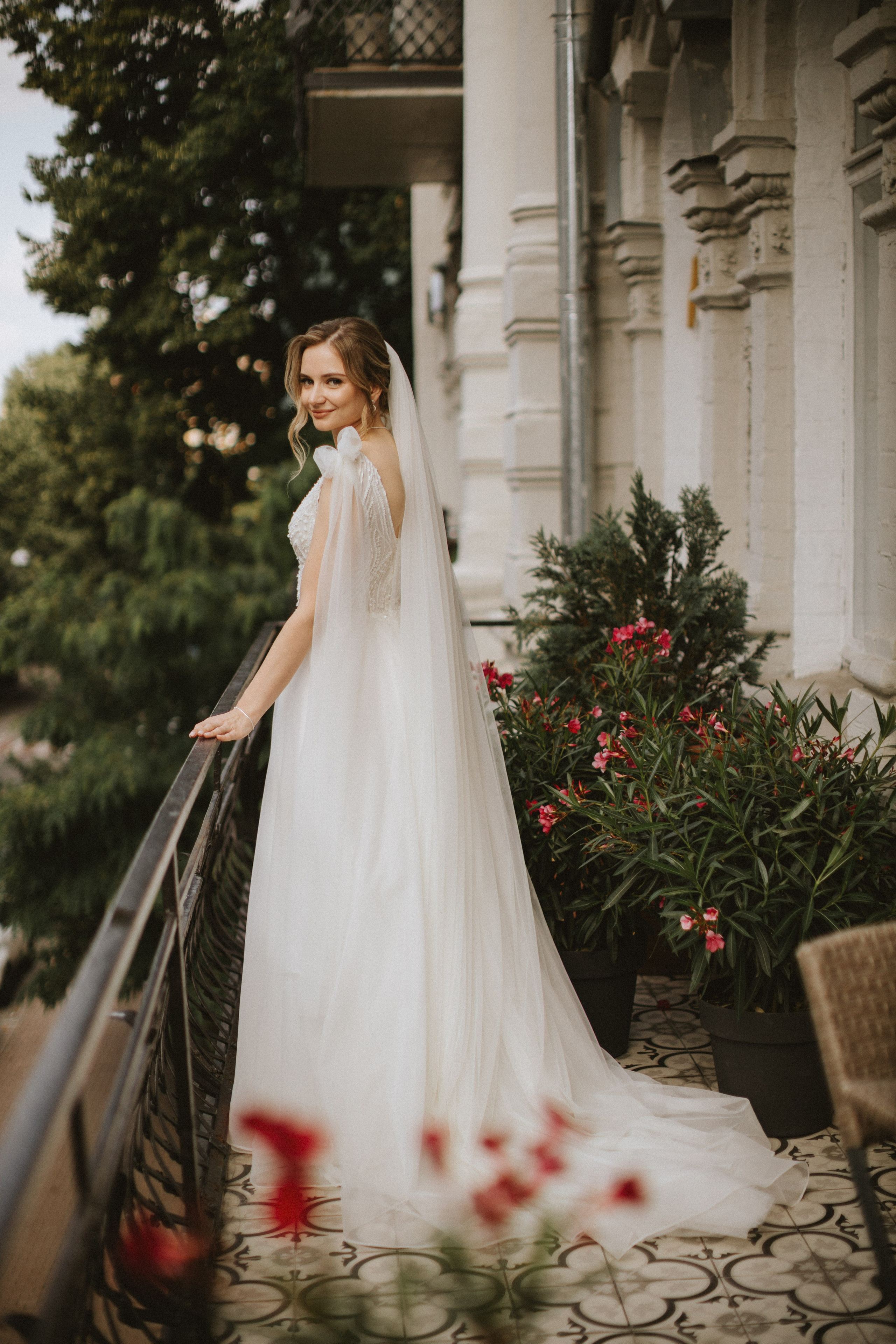 “Una sposa in piedi su un balcone, che sorride con grazia e si guarda alle spalle. Indossa un delicato abito da sposa dal tessuto fluente e un lungo velo. Lo sfondo è caratterizzato da alberi e piante verdi, che esaltano l'atmosfera naturale e romantica del momento”.