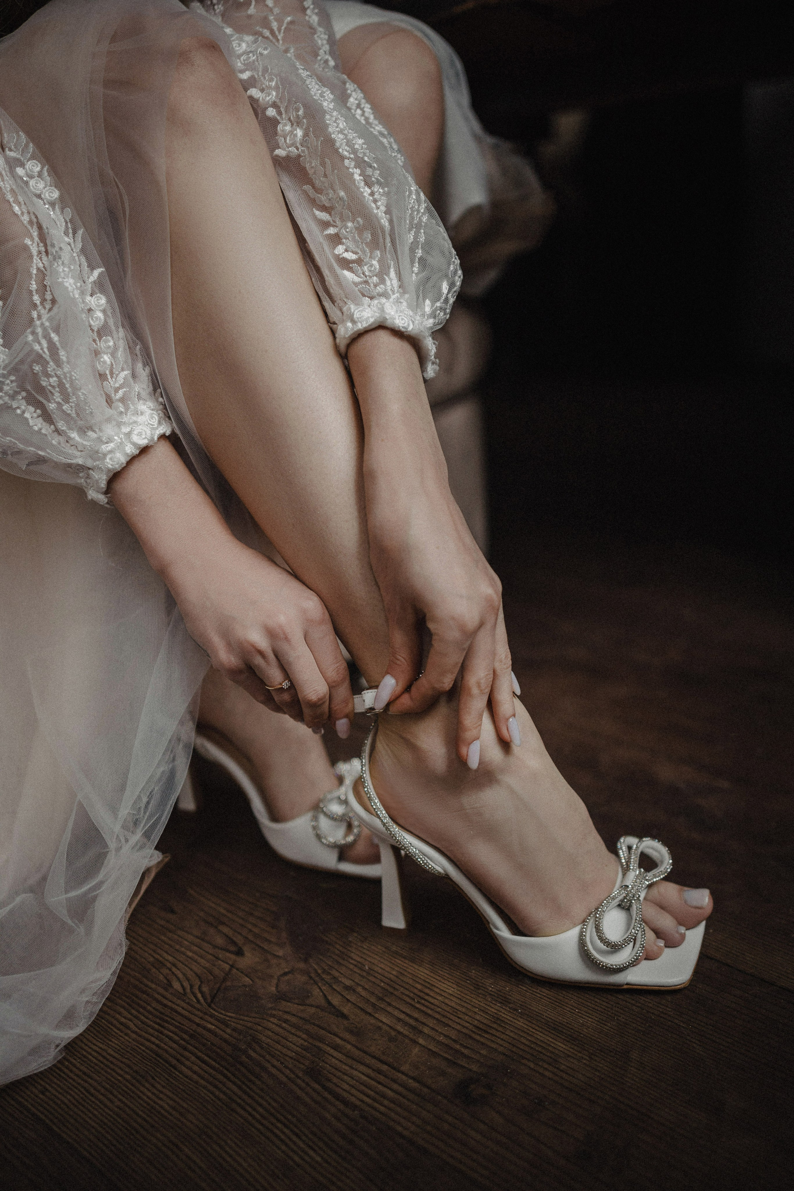 “Un primo piano di una sposa seduta che si aggiusta delicatamente le scarpe bianche con il tacco alto. Le scarpe hanno un delicato dettaglio di fiocco scintillante. Le sue gambe sono graziosamente incrociate e le sue mani stanno aggiustando il cinturino di una scarpa, con una morbida messa a fuoco sulle sue eleganti unghie curate. Indossa una vestaglia da sposa leggera e trasparente con intricati dettagli in pizzo”.