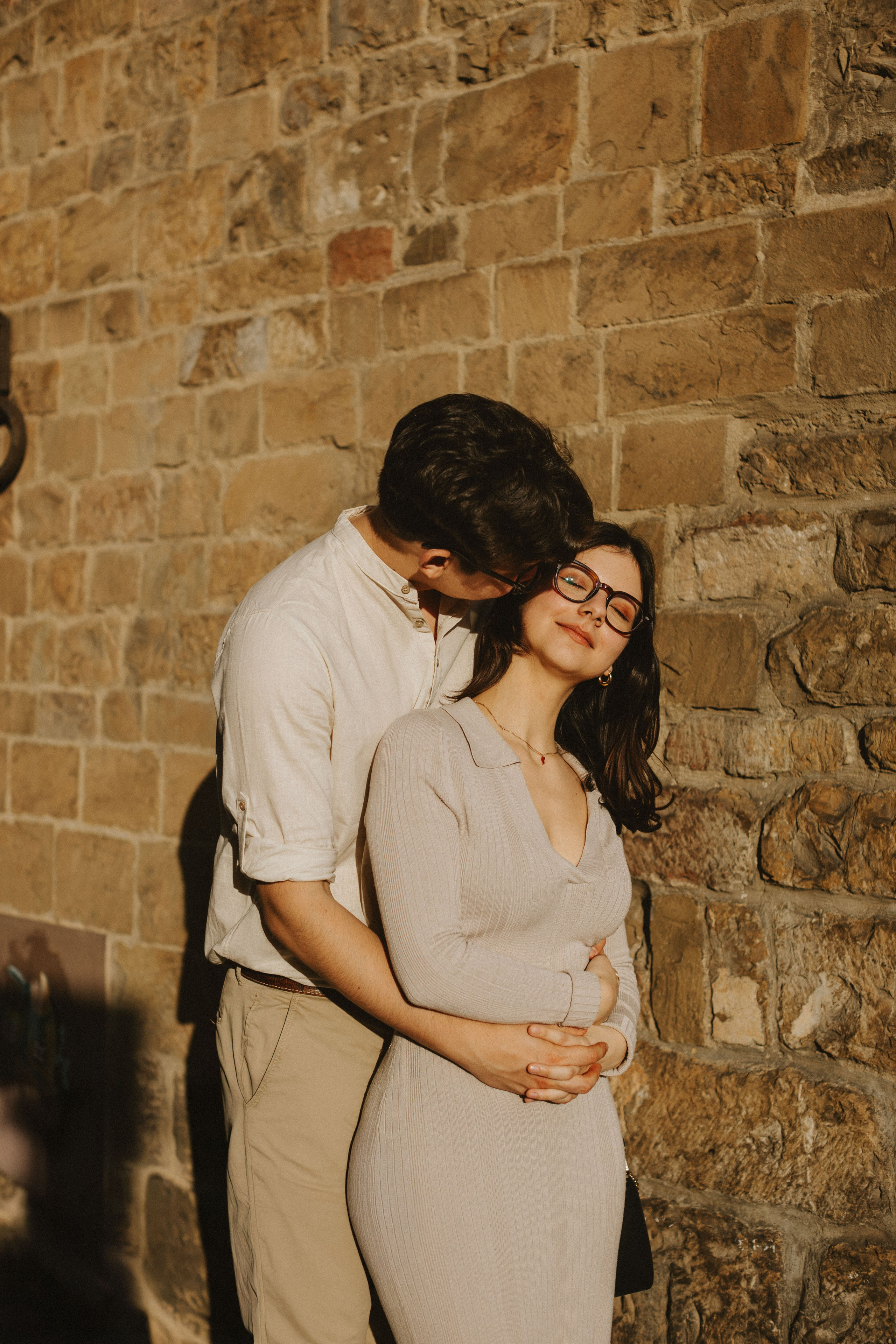 “Una coppia che condivide un momento di tenerezza, sorridendo e abbracciandosi contro un muro di pietra alla luce del sole”.