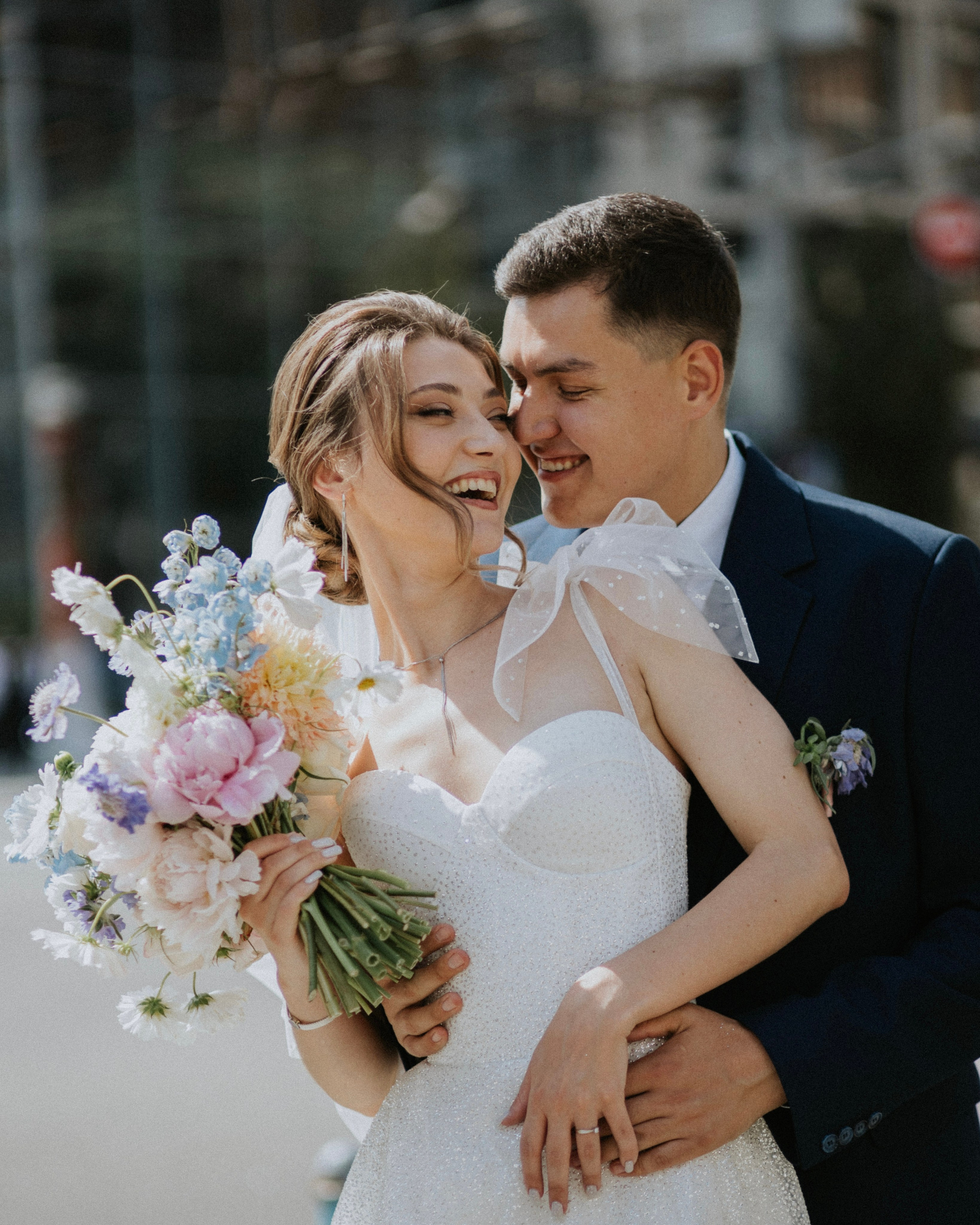 “Un gioioso momento di matrimonio catturato all'aperto mentre gli sposi condividono un abbraccio affettuoso. La sposa indossa un abito bianco scintillante ornato da un delicato fiocco sulla spalla e tiene in mano un colorato bouquet di peonie, fiori di campo e verde. Lo sposo, vestito con un abito scuro, abbraccia amorevolmente la sposa mentre si sorridono l'un l'altro, con la morbida luce del sole che proietta una calda luce sui loro volti. Lo sfondo mostra un accenno di una vivace scena cittadina”.