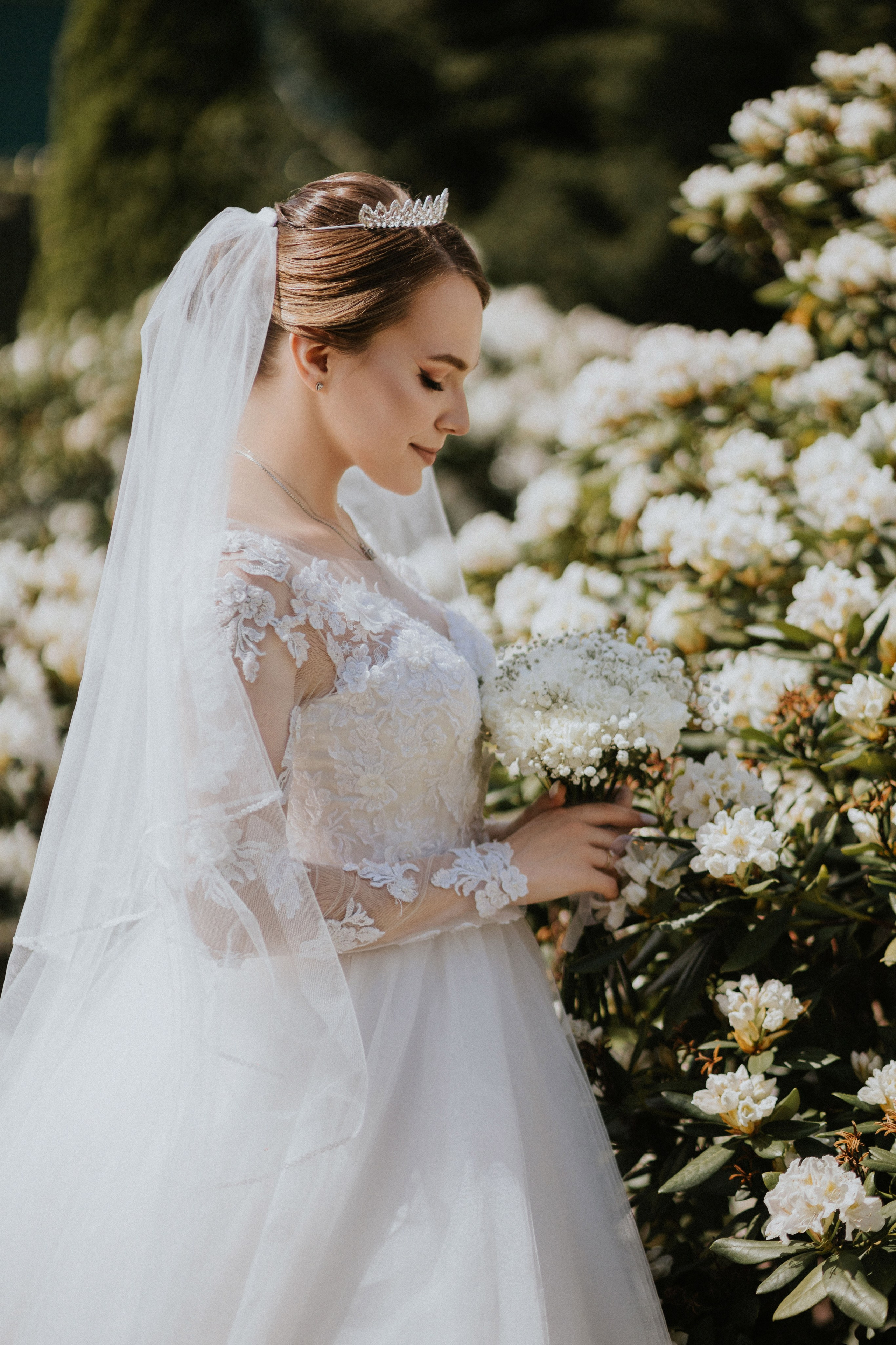 “Una sposa si trova in mezzo a un mare di fiori bianchi, tenendo delicatamente in mano un bouquet di delicati fiori bianchi. Ha un'espressione dolce sul viso, con gli occhi chiusi, e la sua testa è adornata da un diadema scintillante. La luce del sole mette in risalto gli intricati dettagli in pizzo del suo abito da sposa e il velo fluente, creando un'atmosfera serena e pacifica.”