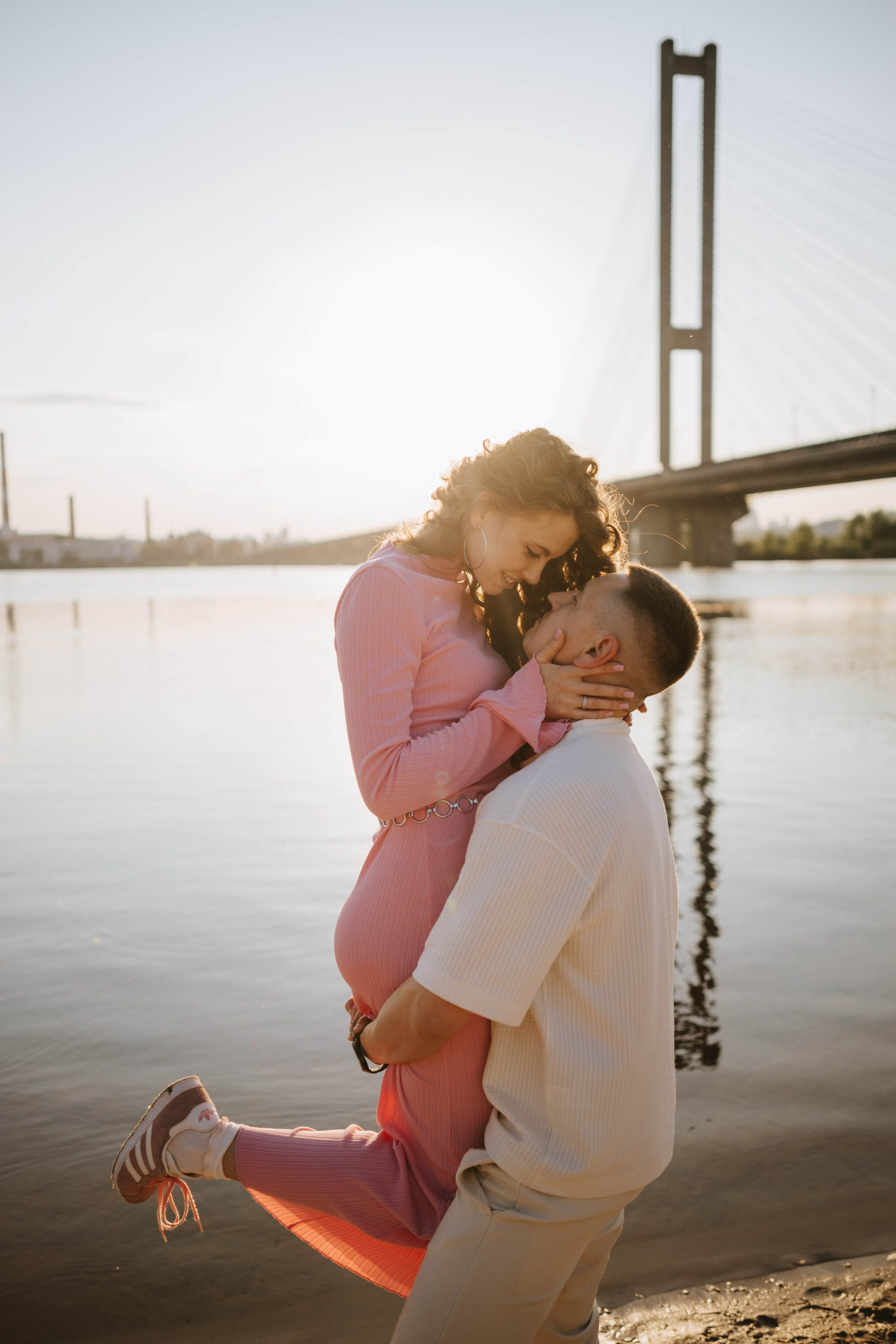 “Una coppia che si gode un momento romantico in riva al mare mentre l'uomo solleva la donna tra le braccia. Il sole al tramonto crea un bagliore dorato, con il ponte industriale sullo sfondo che aggiunge un'atmosfera bella e intima alla scena.”