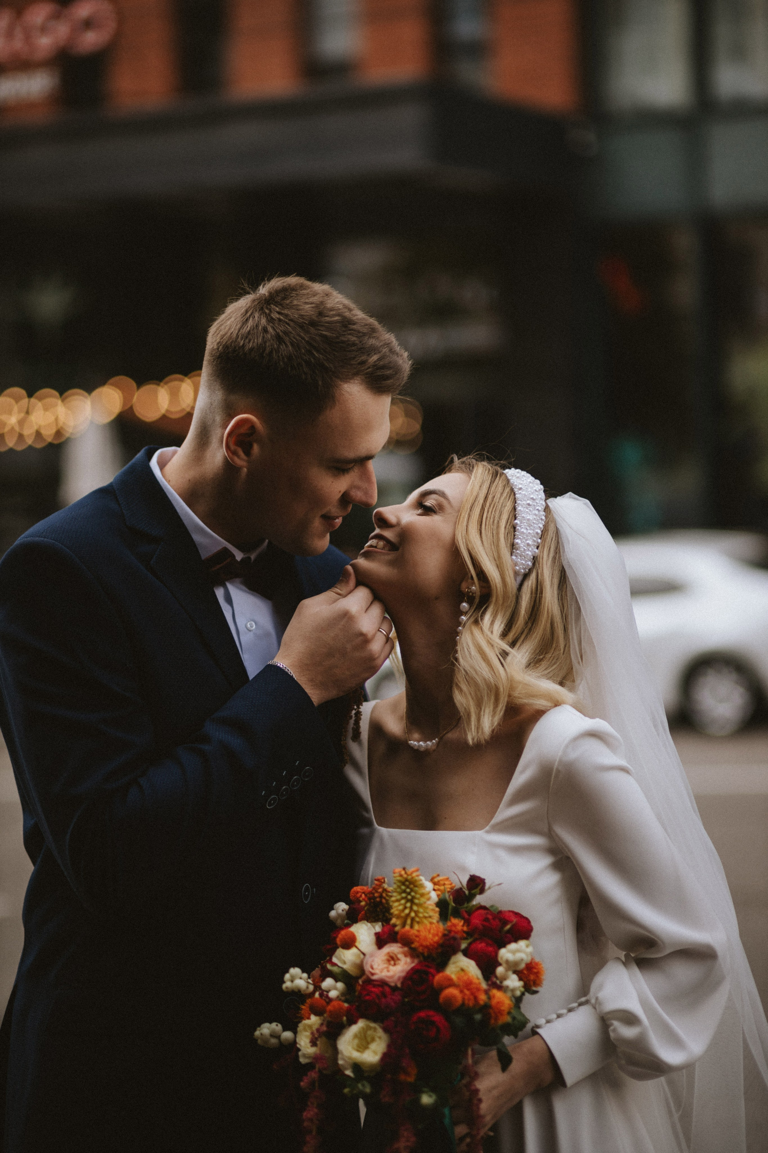 “Una sposa e uno sposo condividono un dolce momento all'aperto. Lo sposo tiene delicatamente il viso della sposa, guardandola amorevolmente mentre lei gli sorride. La sposa tiene in mano un vivace bouquet di fiori rossi, arancioni e gialli e indossa un delicato cerchietto di perle con i suoi capelli biondi e ondulati. Lo sposo è vestito con un abito blu scuro e un papillon. Lo sfondo è caratterizzato da edifici cittadini sfocati e luci soffuse, che creano un'atmosfera romantica”.