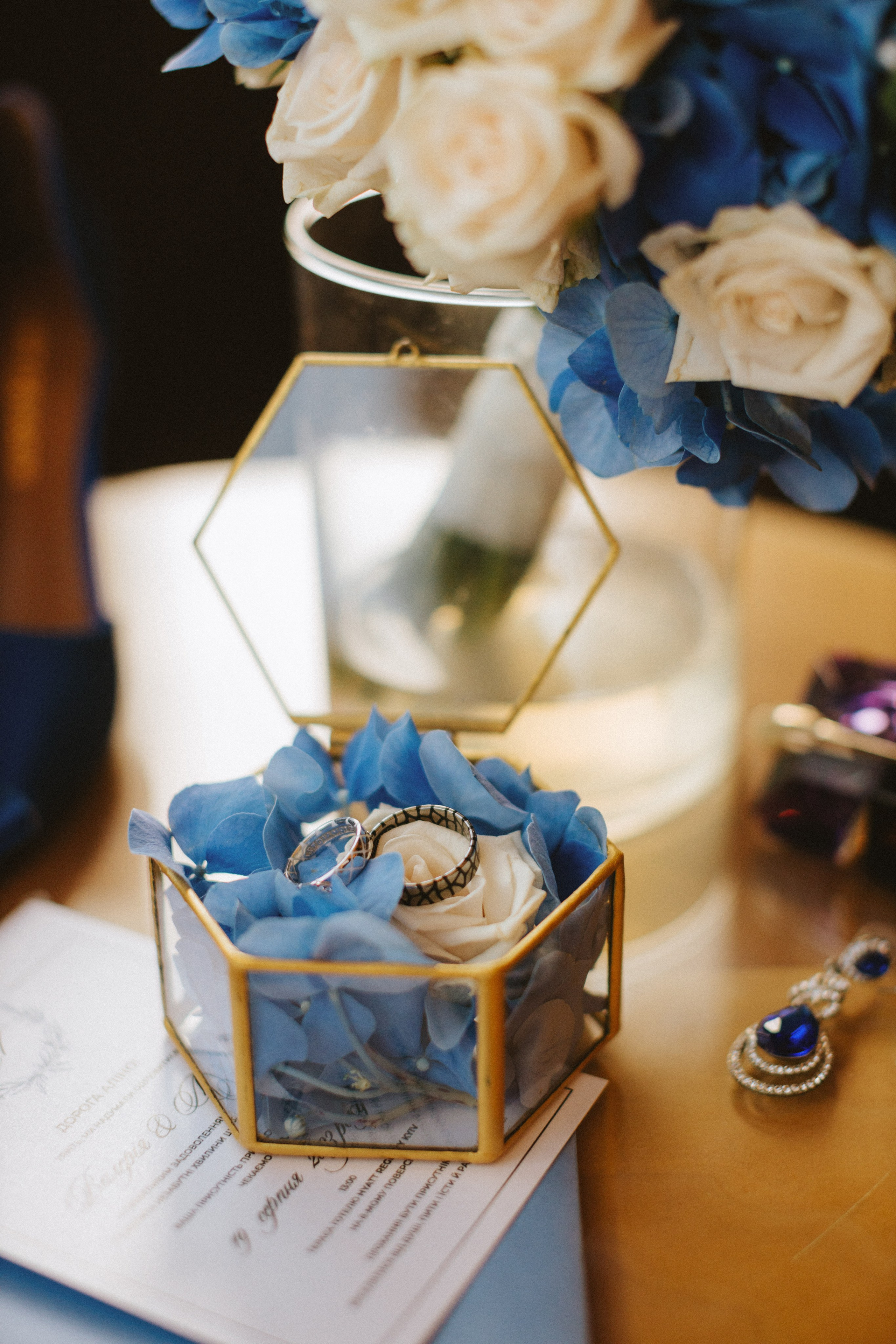 “Un'elegante esposizione di fedi nuziali collocate in una scatola di vetro riempita di morbidi petali blu e di una rosa bianca. Gli anelli sono esposti sullo sfondo di un delicato bouquet e di una partecipazione di nozze, con un gioiello scintillante in un angolo”.