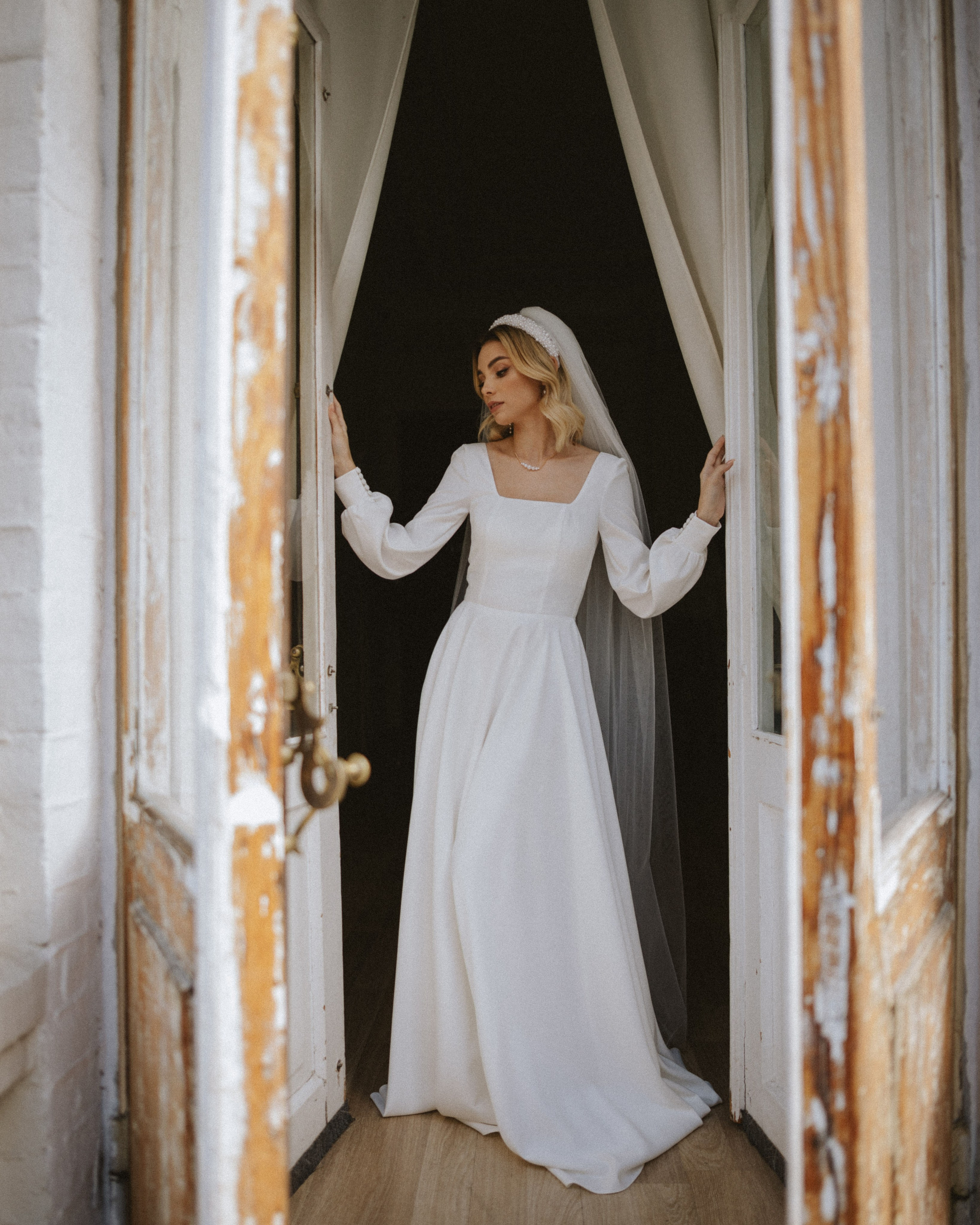 “The bride in a wedding dress and veil stands by the door, with a gentle smile, preparing for the wedding.”