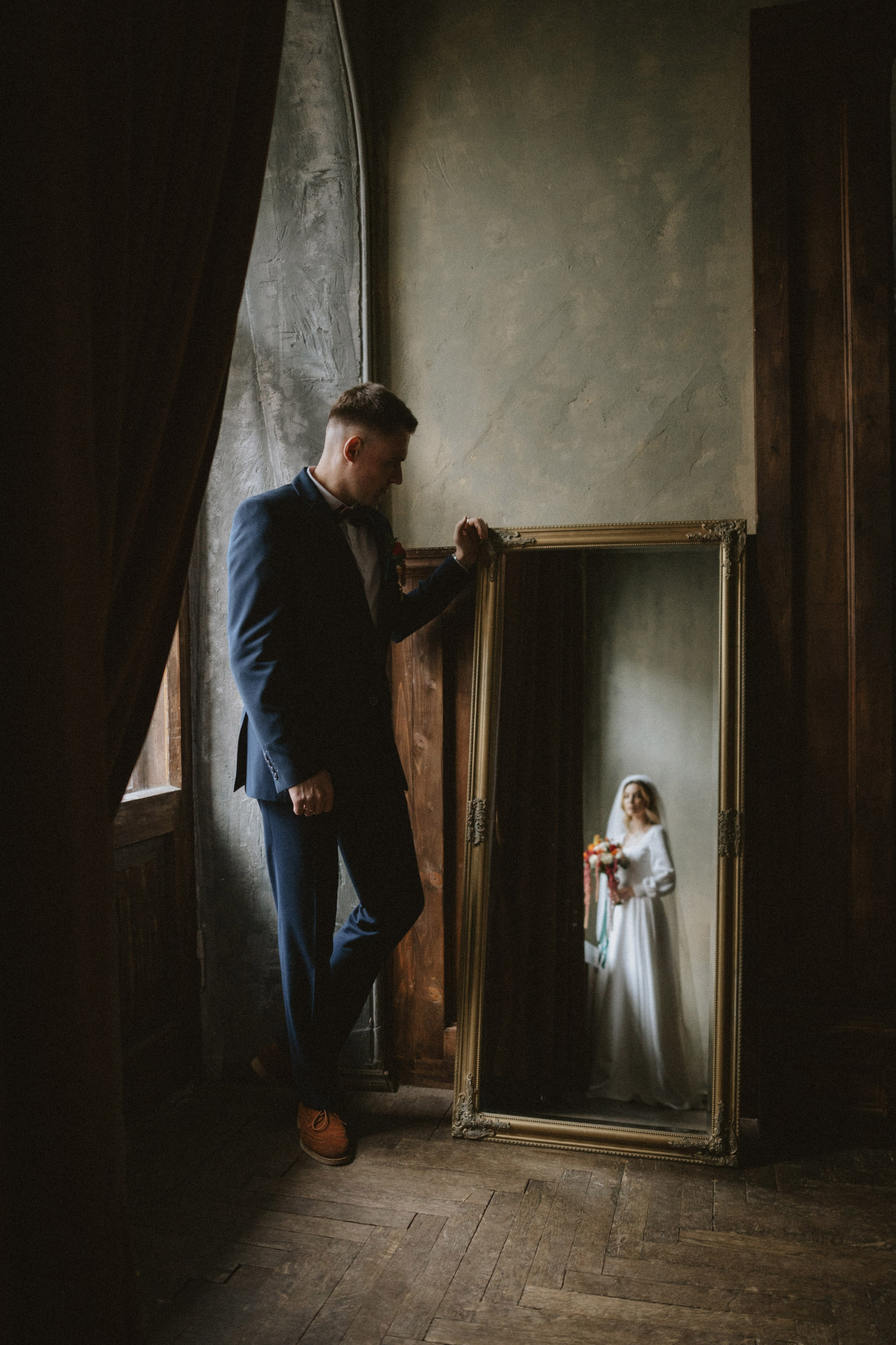 “Uno sposo è in piedi davanti a uno specchio, con la mano delicatamente appoggiata alla cornice, mentre guarda il riflesso della sua sposa in abito da sposa, con in mano un bouquet. L'illuminazione calda e intima della stanza crea un'atmosfera nostalgica e romantica”.