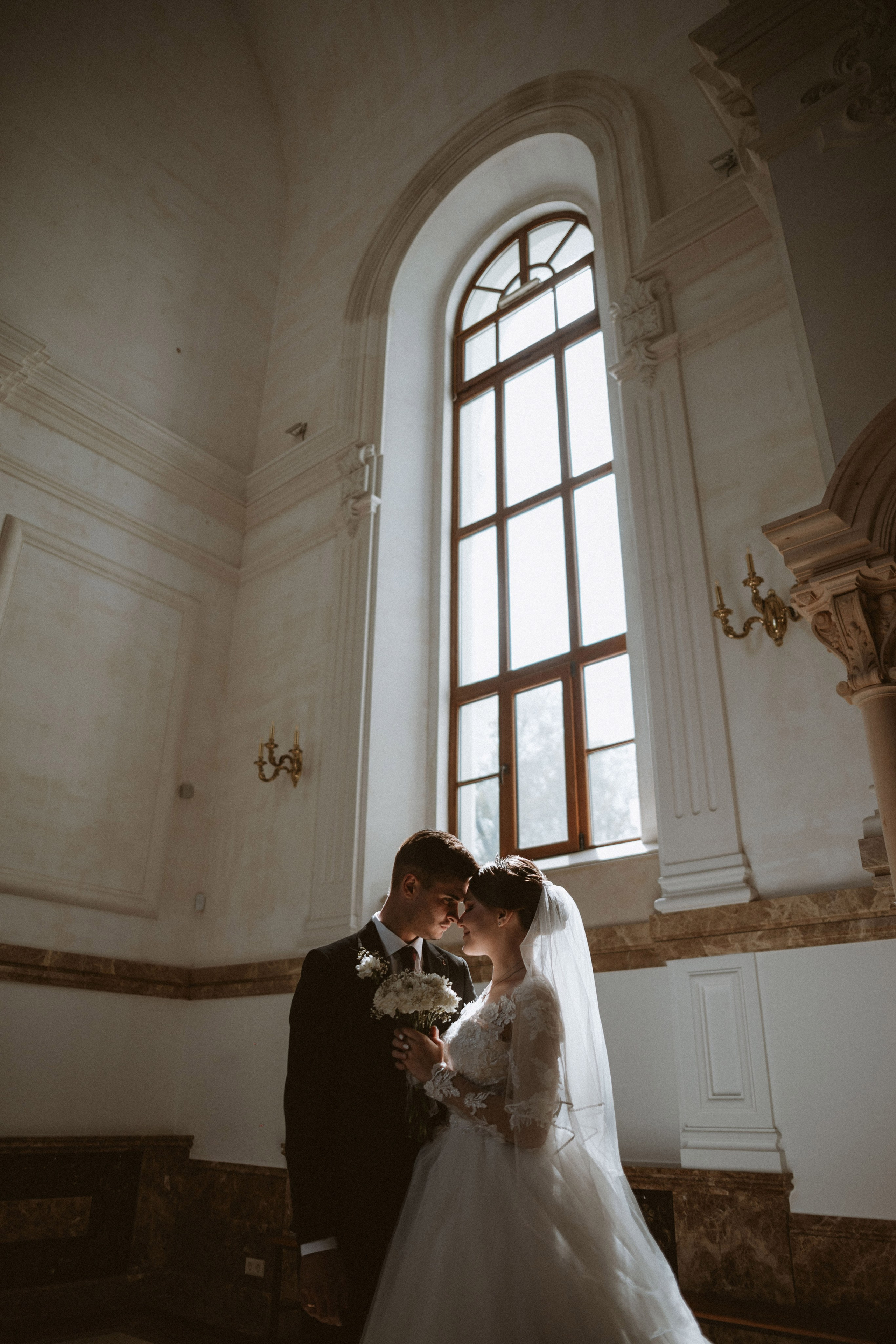 “Romantica foto di matrimonio di una coppia che condivide un momento di tenerezza al chiuso, con la morbida luce del sole che filtra da un'ampia finestra. La sposa, con un bouquet di fiori in mano, indossa un elegante abito da sposa in pizzo e un velo. Lo sposo, in un classico abito nero, si china per un abbraccio stretto”.