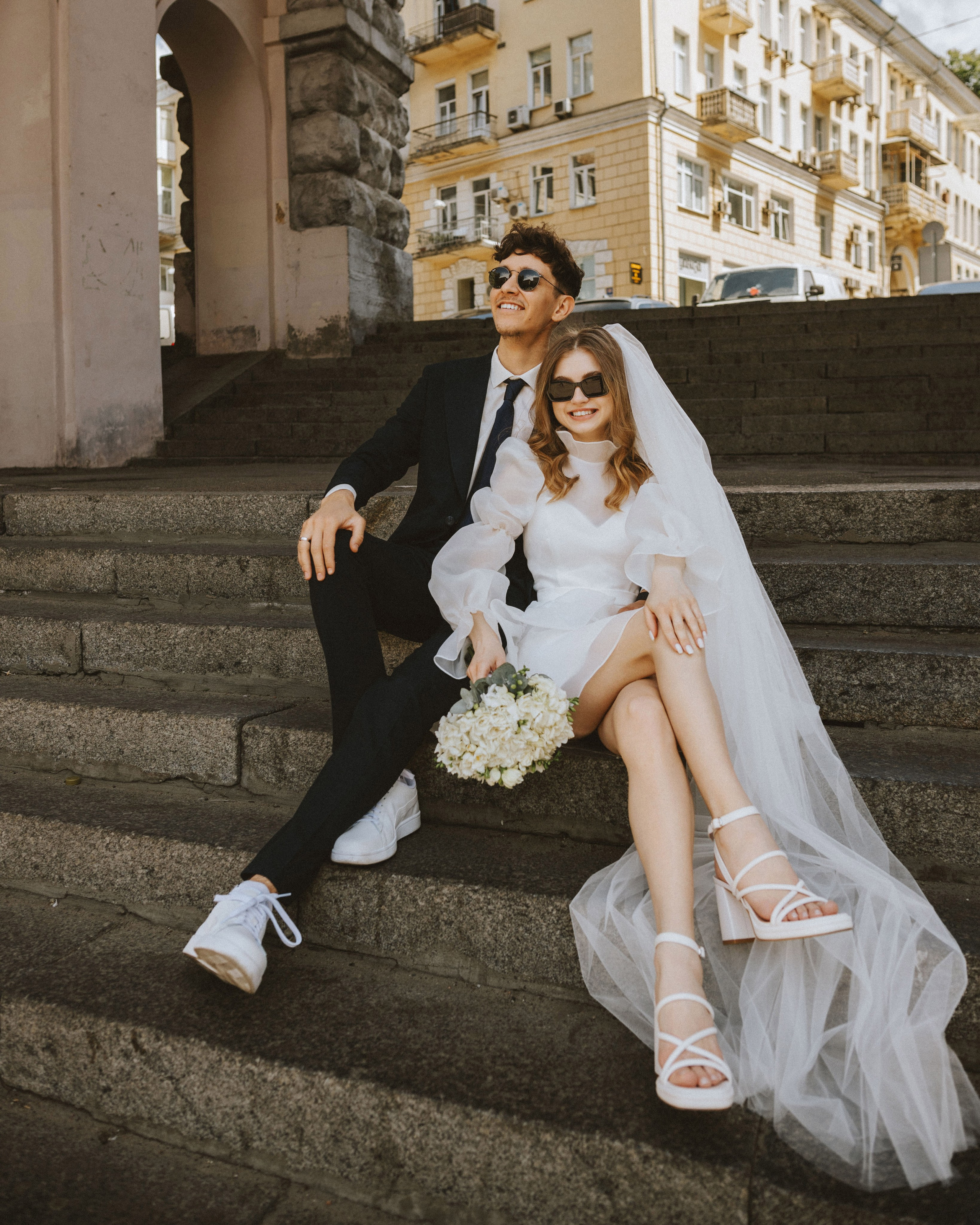 “Una coppia felice seduta sulle scale, entrambi con gli occhiali da sole. La sposa indossa un elegante abito da sposa bianco e un velo, tenendo in mano un bouquet, mentre lo sposo è vestito con un abito nero e scarpe da ginnastica bianche. Entrambi hanno un aspetto rilassato e gioioso in questo ambiente moderno e urbano”.