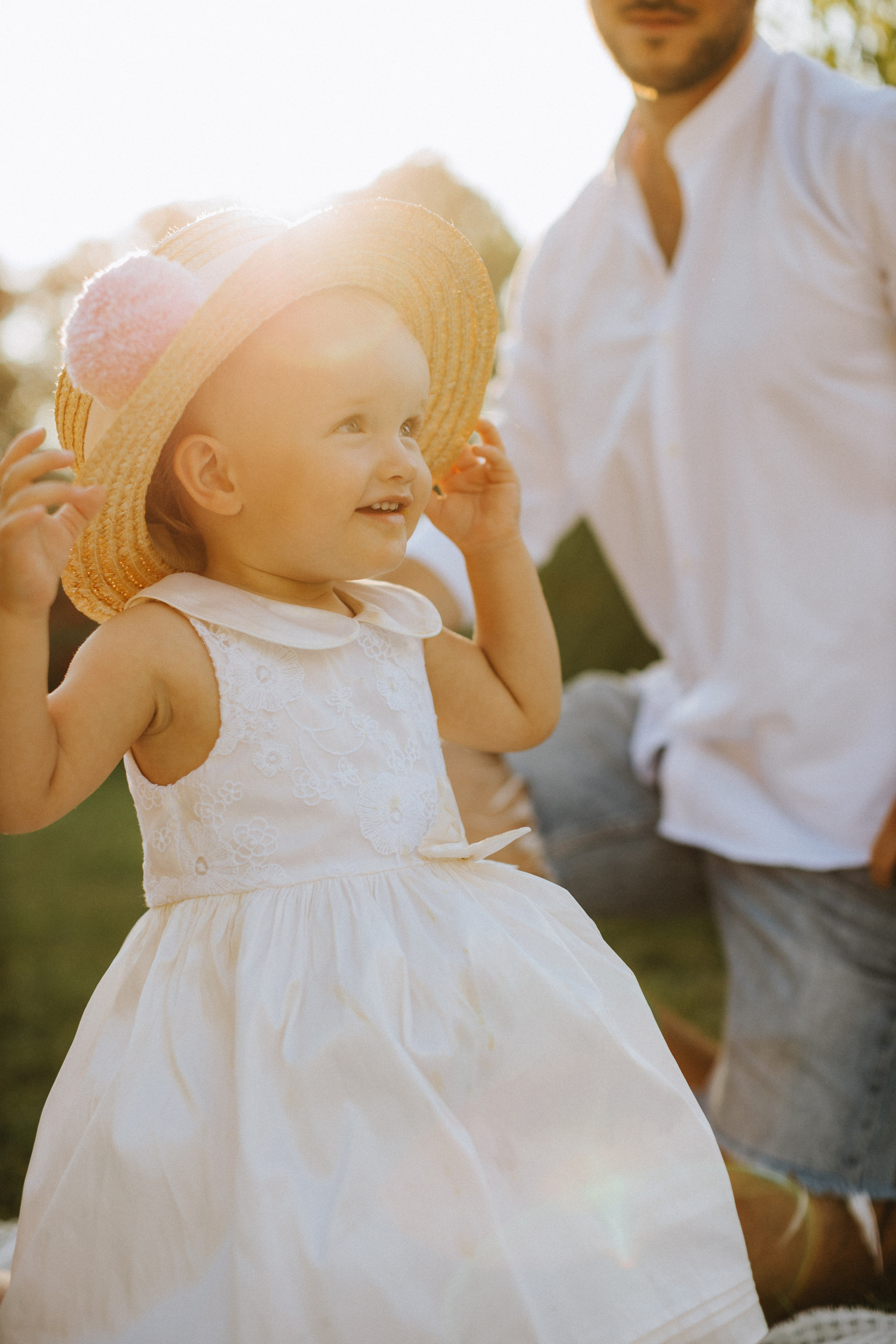 “Una bambina gioiosa che indossa un vestito bianco e un cappello di paglia con un pompon e sorride mentre si aggiusta giocosamente il cappello. Il sole crea uno sfondo morbido e luminoso e sullo sfondo si vede il padre, creando un momento caldo e felice”.