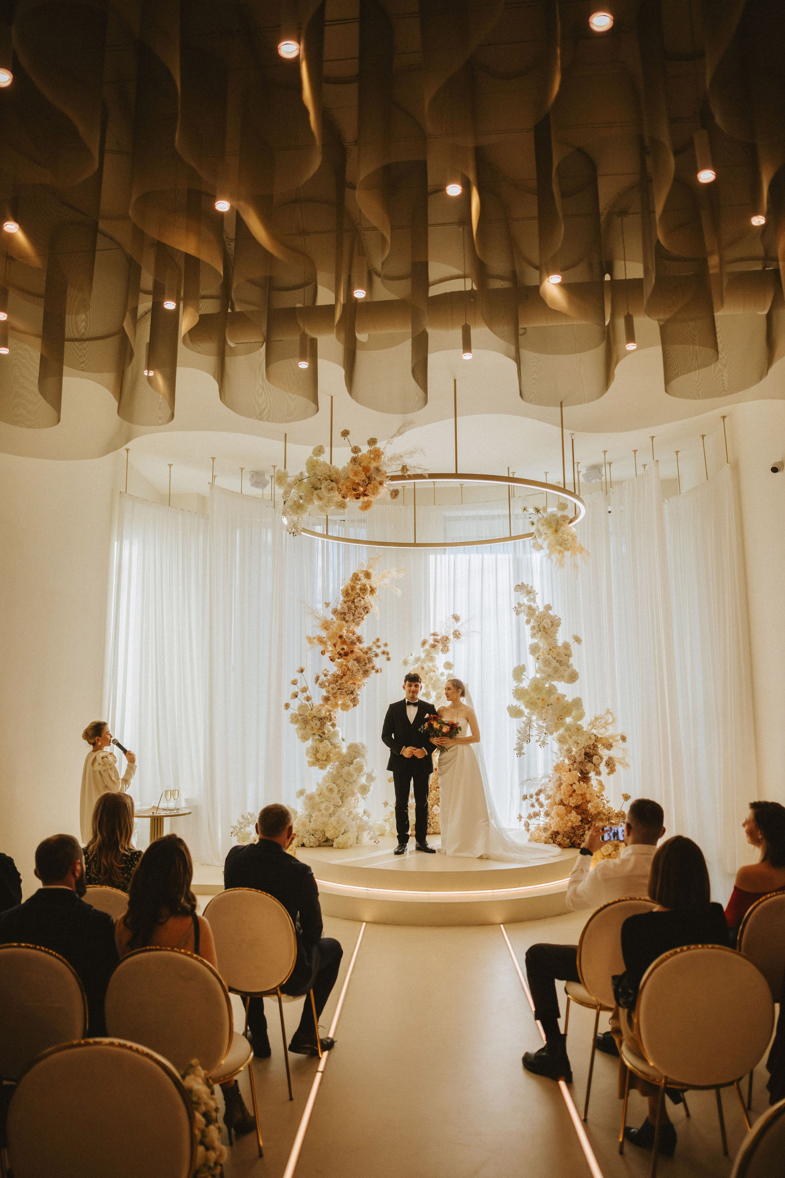 “Una coppia in piedi sul palco della cerimonia nuziale, circondata da composizioni floreali. La sposa in abito bianco regge il bouquet, accanto allo sposo in smoking nero, mentre l'officiante parla”.