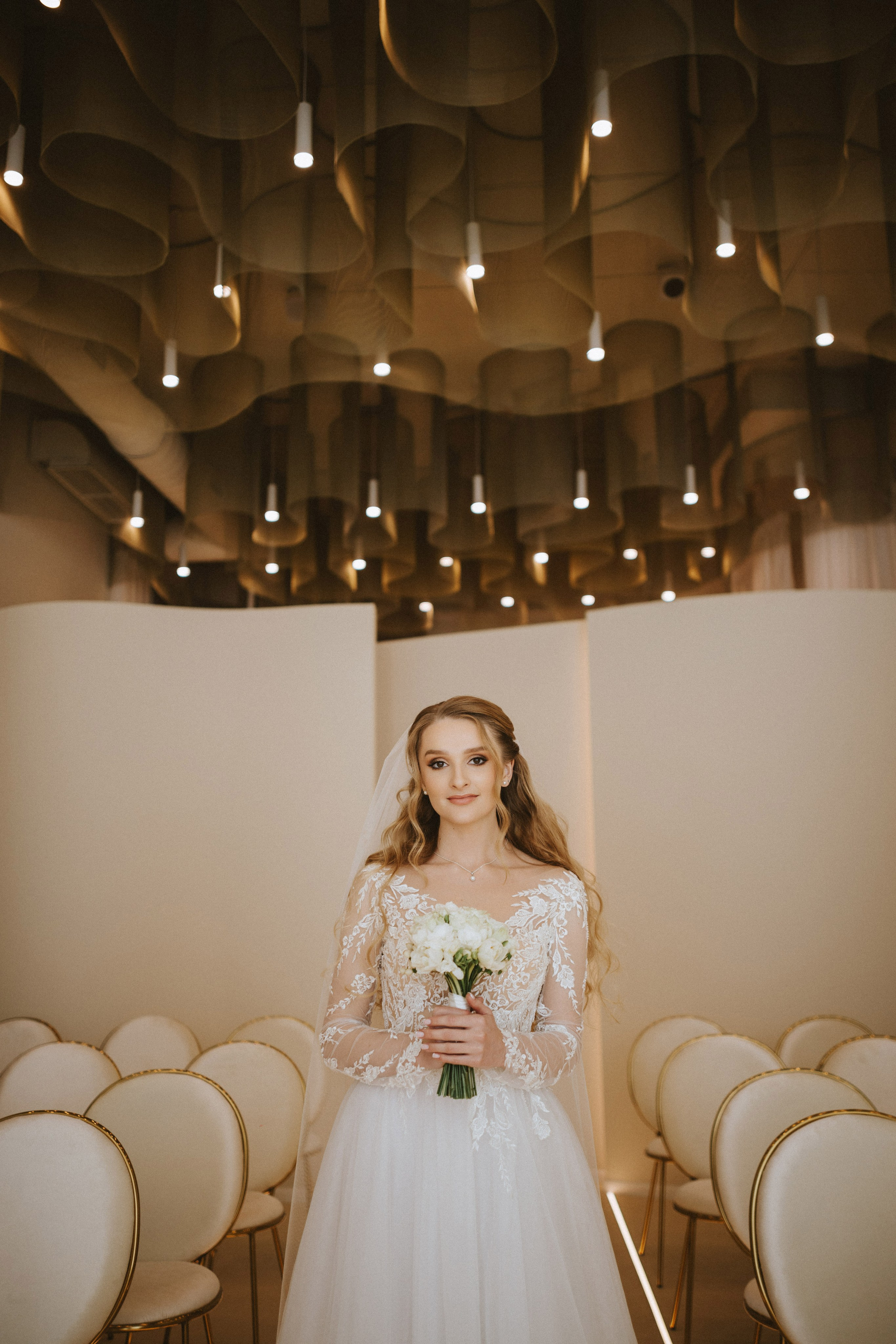 “Una sposa si trova al centro di una moderna location per matrimoni, sorridendo calorosamente mentre regge il suo bouquet di fiori bianchi. L'interno elegante è caratterizzato da luci morbide e curve sul soffitto e da decorazioni minimaliste, che creano un'atmosfera serena ed elegante. Il suo abito da sposa, con delicati dettagli in pizzo, completa la raffinata eleganza dello spazio”.
