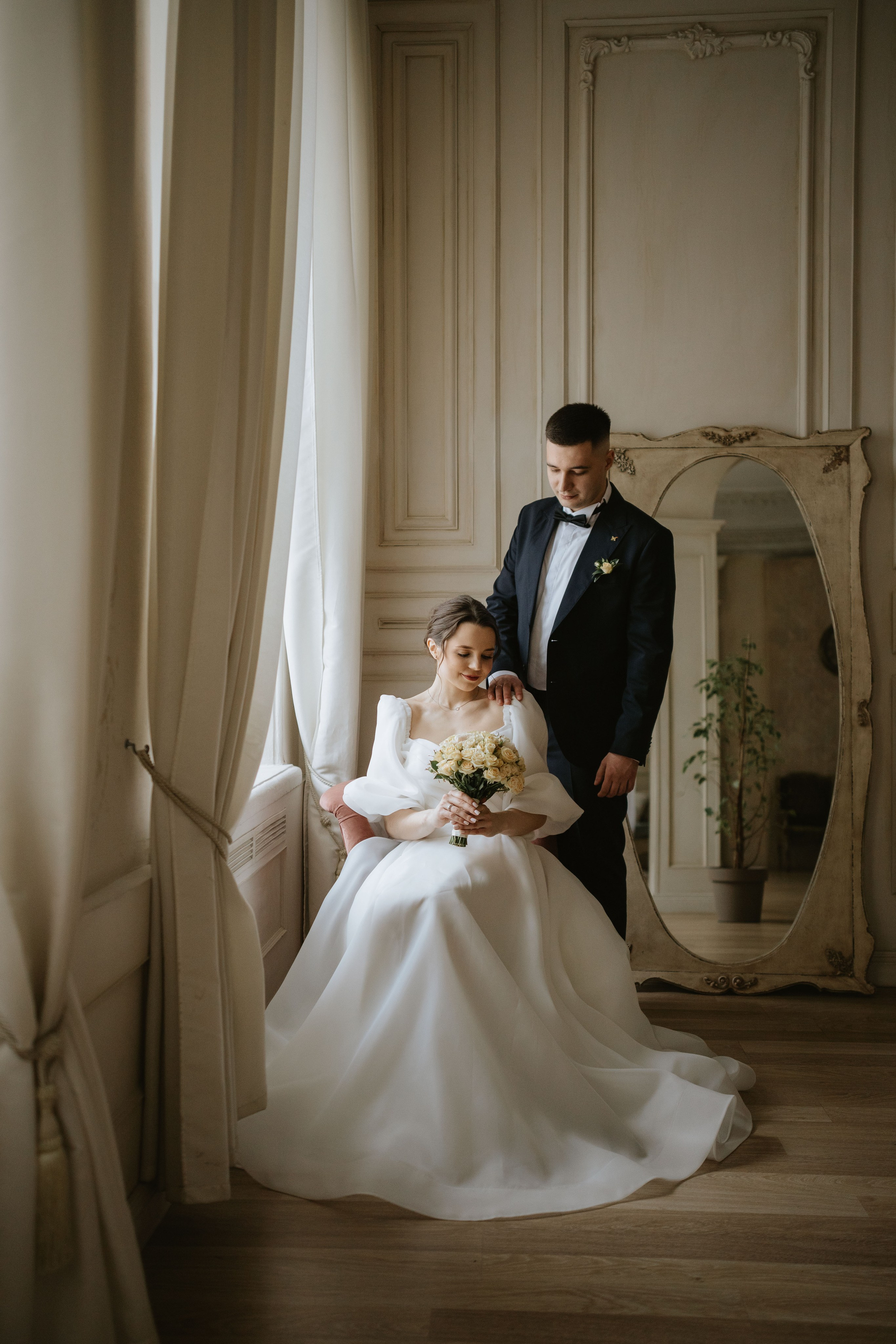 “Una sposa in un bellissimo abito bianco siede con grazia su una sedia rosa, tenendo in mano un bouquet di rose. Lo sposo è in piedi dietro di lei e le appoggia delicatamente la mano sulla spalla, guardandola con affetto. L'ambiente è una stanza elegante e luminosa con grandi finestre e uno specchio decorativo sullo sfondo”.