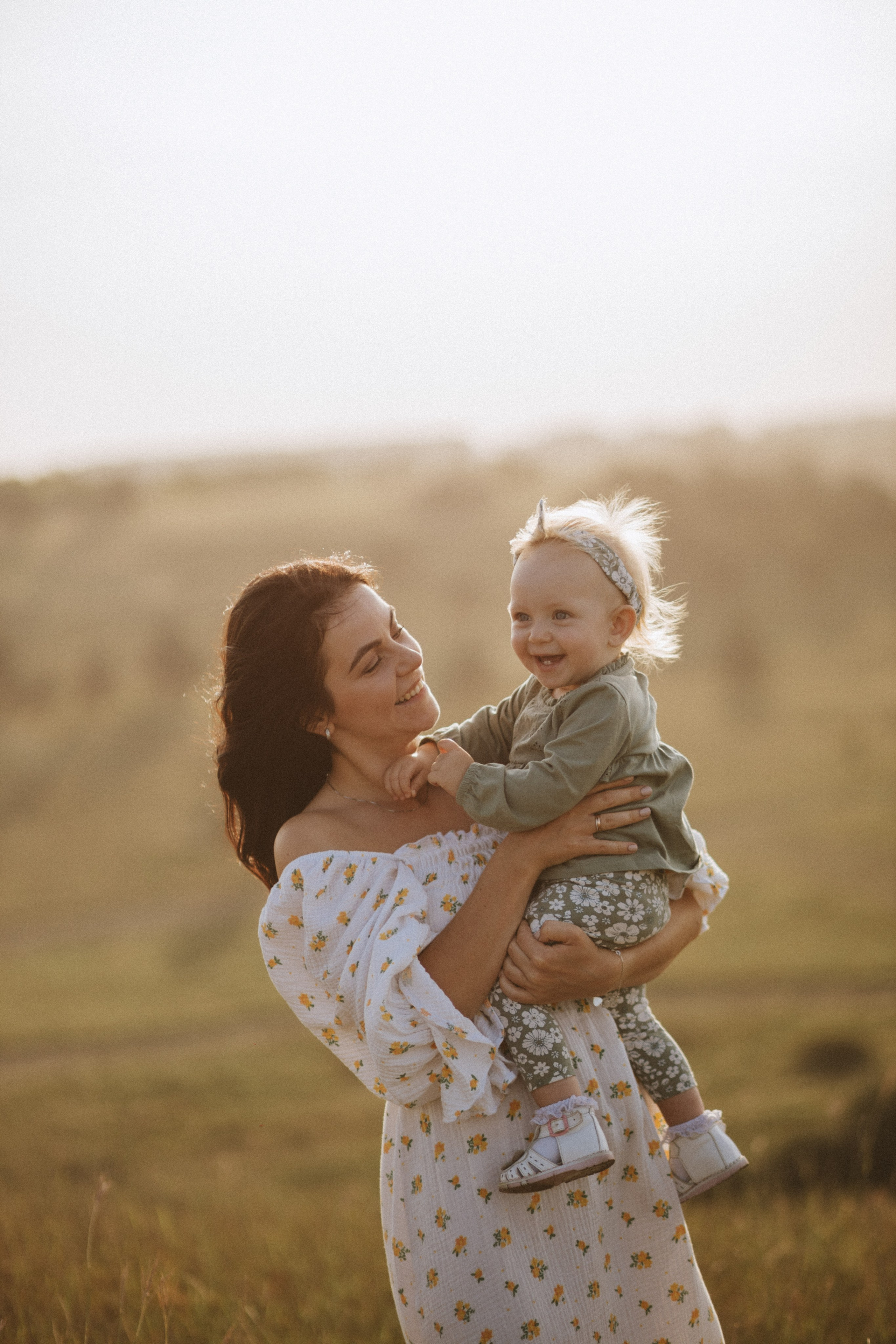 “Una madre gioiosa tiene in braccio il suo bambino sorridente, entrambi emanano calore e amore. La madre indossa un abito bianco senza spalline con un motivo floreale, mentre il bambino è vestito con un completo verde con leggings floreali e una fascia per capelli. La luce dorata del sole mette in risalto i loro volti, con uno sfondo morbido e sfocato di un campo erboso, creando un momento di pace e affetto tra loro”.