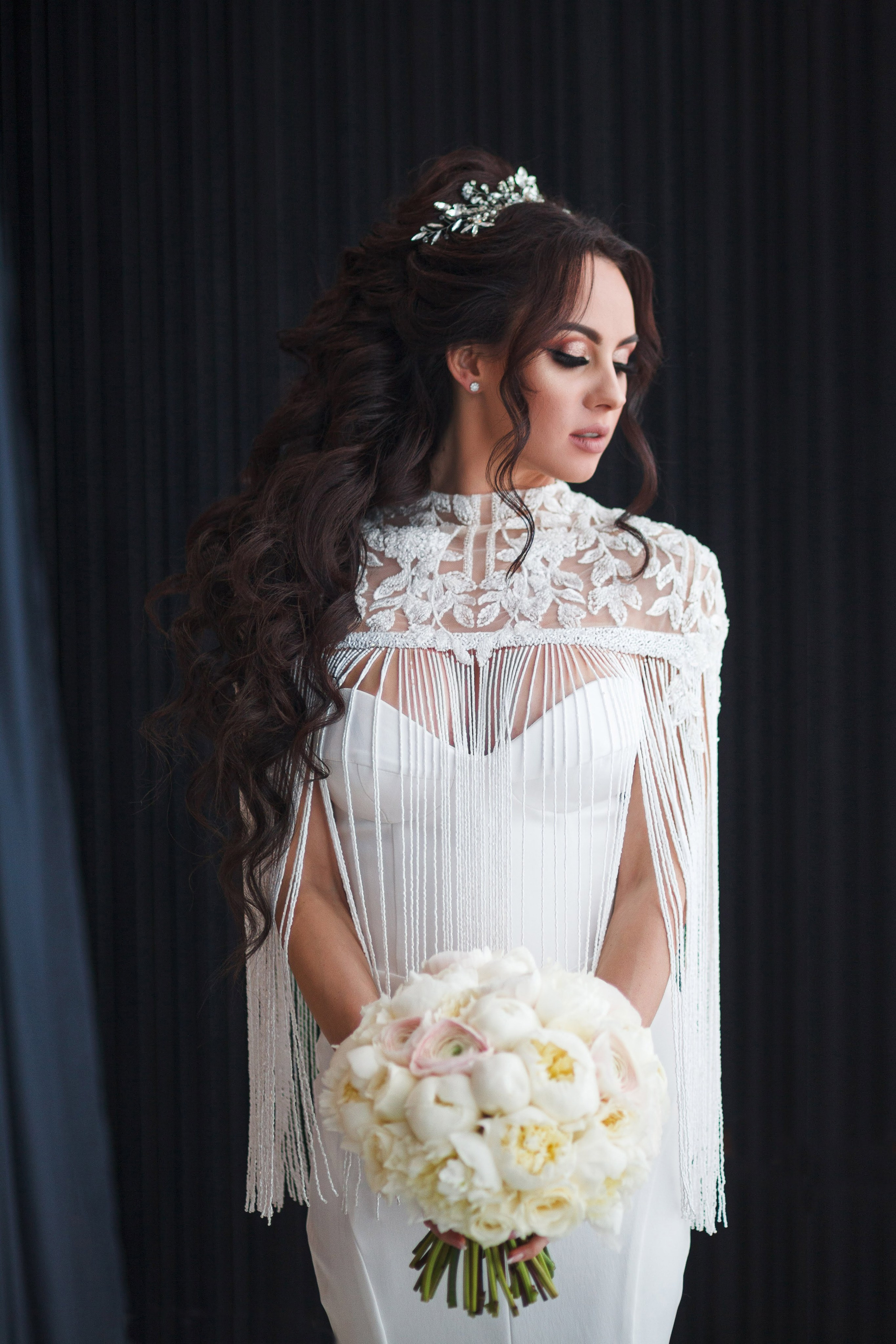 “Una sposa in uno splendido abito da sposa con un delicato dettaglio di frange sulla scollatura, con in mano un bouquet di peonie bianche. I suoi lunghi e voluminosi ricci sono splendidamente acconciati e lei indossa un accessorio per capelli scintillante, creando un look elegante e senza tempo per il suo giorno speciale.”