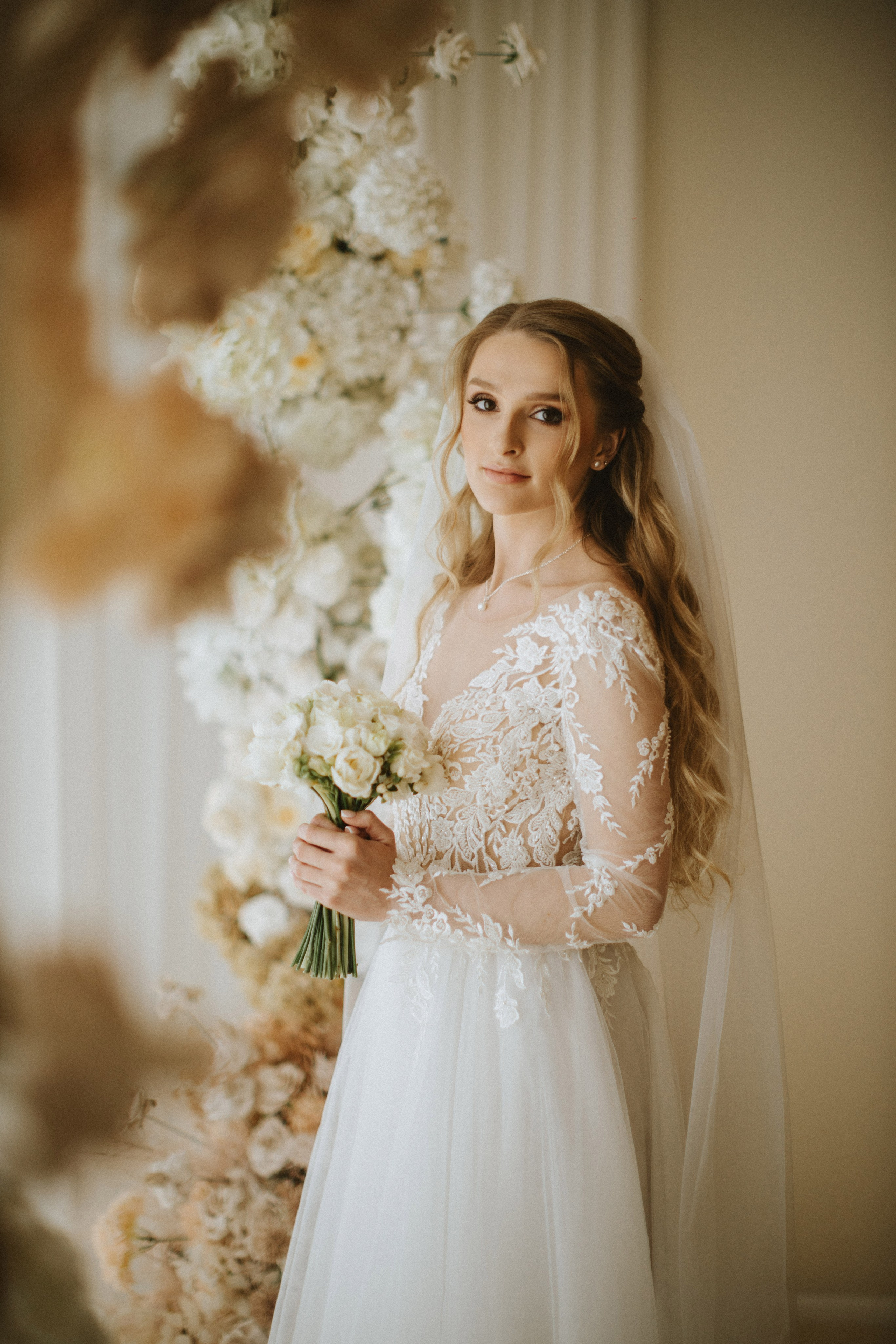 “Una sposa si erge con grazia, tenendo in mano un bouquet di fiori bianchi. Ha un'espressione dolce e serena mentre guarda l'obiettivo, i capelli acconciati a onde sciolte e il velo che le scorre dietro. Gli intricati dettagli in pizzo dell'abito a maniche lunghe aggiungono un tocco di eleganza. Lo sfondo floreale sfuma dolcemente sullo sfondo, esaltando l'atmosfera romantica del momento”.