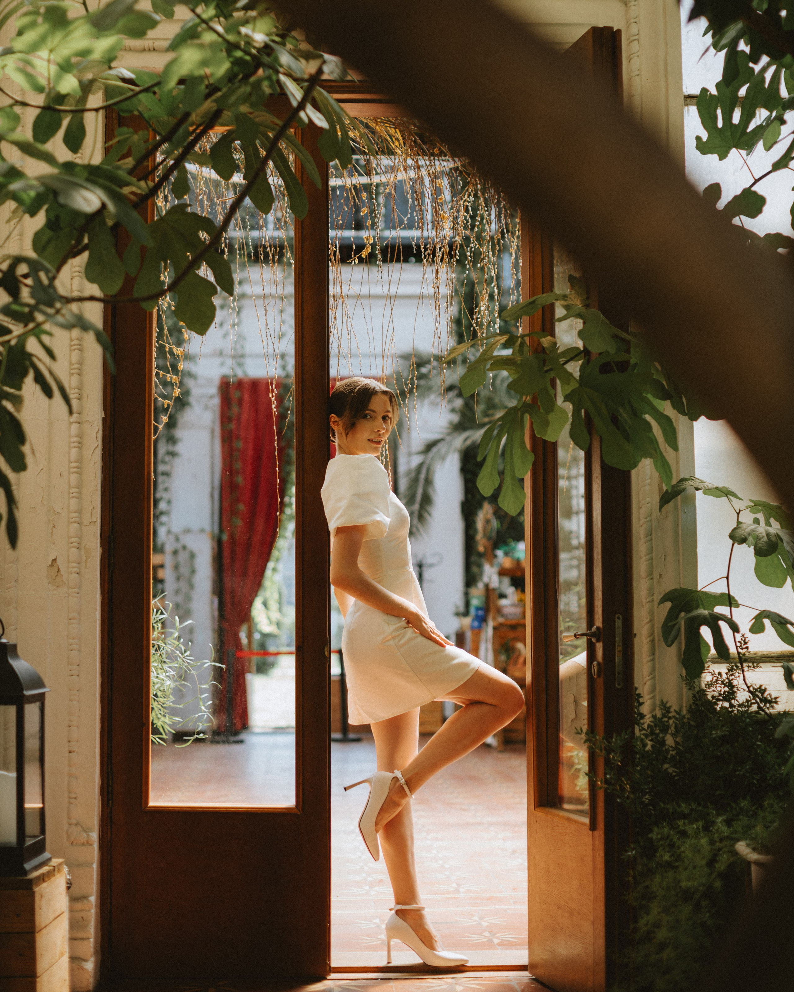 “The bride in a stylish short dress and white heels stands in a doorway, surrounded by greenery.”